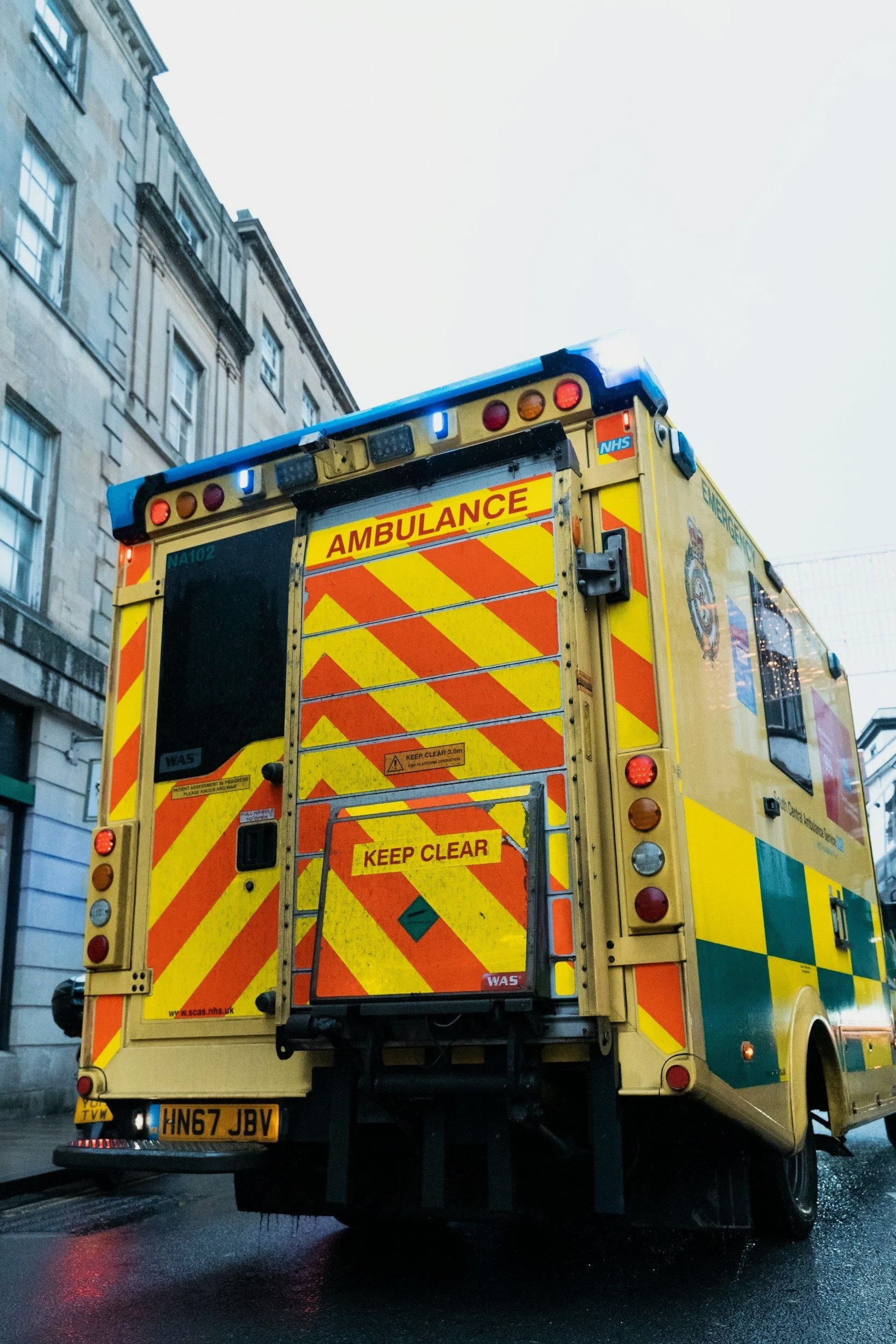 Rear view of a yellow ambulance with red and yellow chevron patterns, emergency lights, and 'KEEP CLEAR' sign, parked on a wet street with tall buildings in the background.
