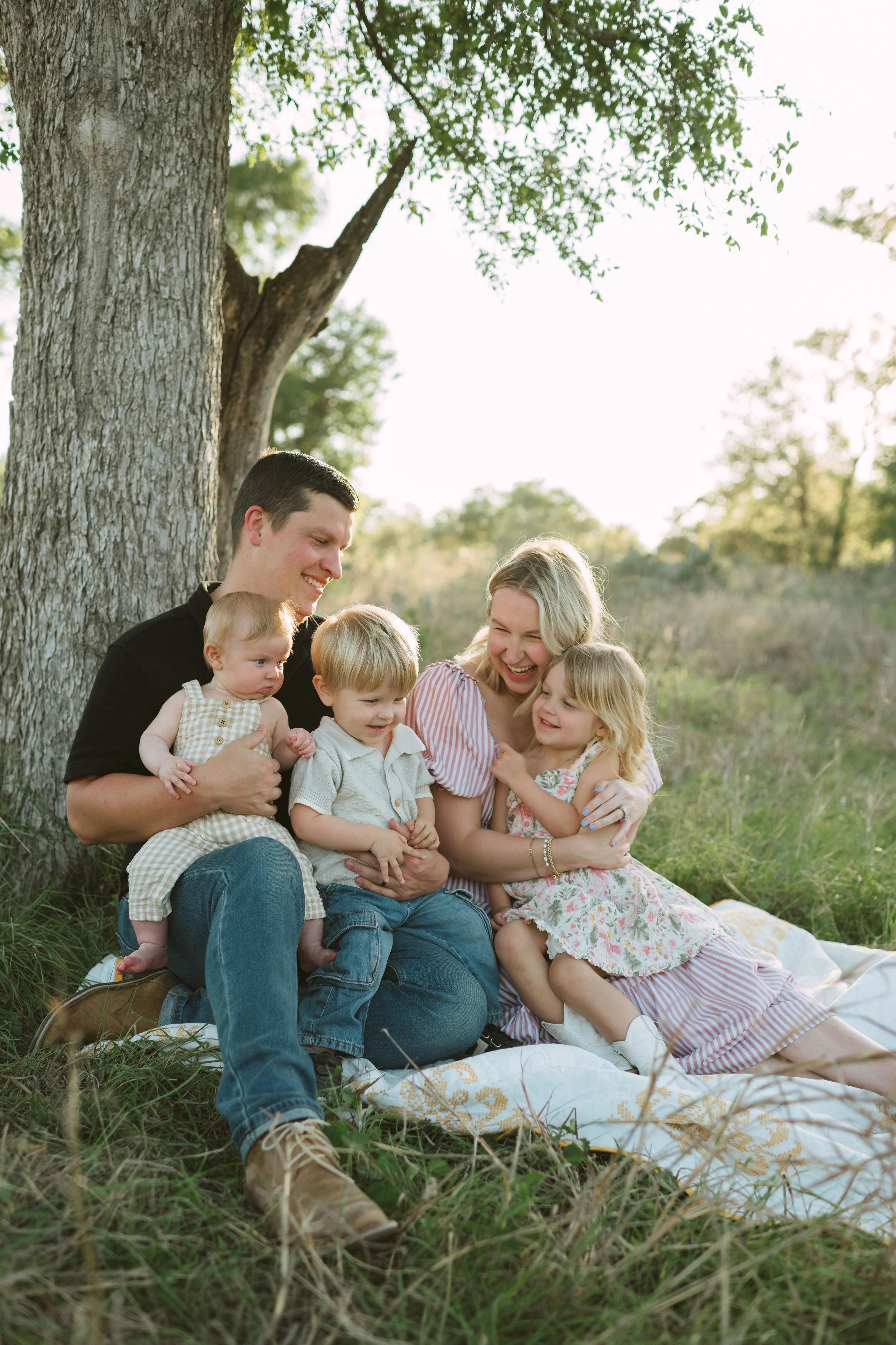 A family of six, including two adults and four children, sitting and cuddling under a large tree in a grassy outdoor setting during the daytime, smiling and enjoying each other's company.