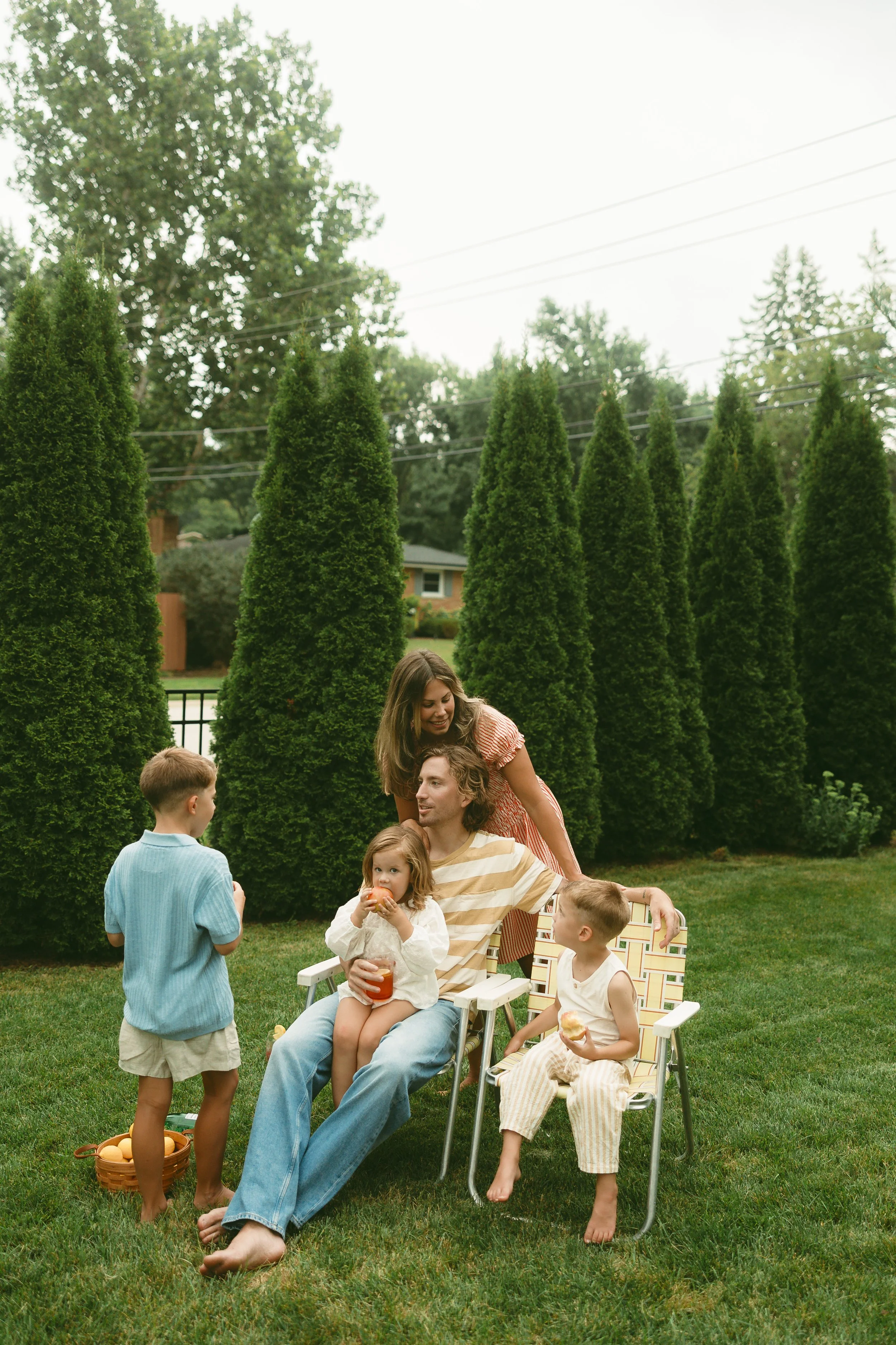A family of six enjoying an outdoor gathering in a backyard with tall green trees and a house in the background. The group includes a man sitting in a white chair, two women, and three children. The man is holding a young girl with curly hair and a drink. The children are talking and eating, and there is a basket of eggs on the grass.