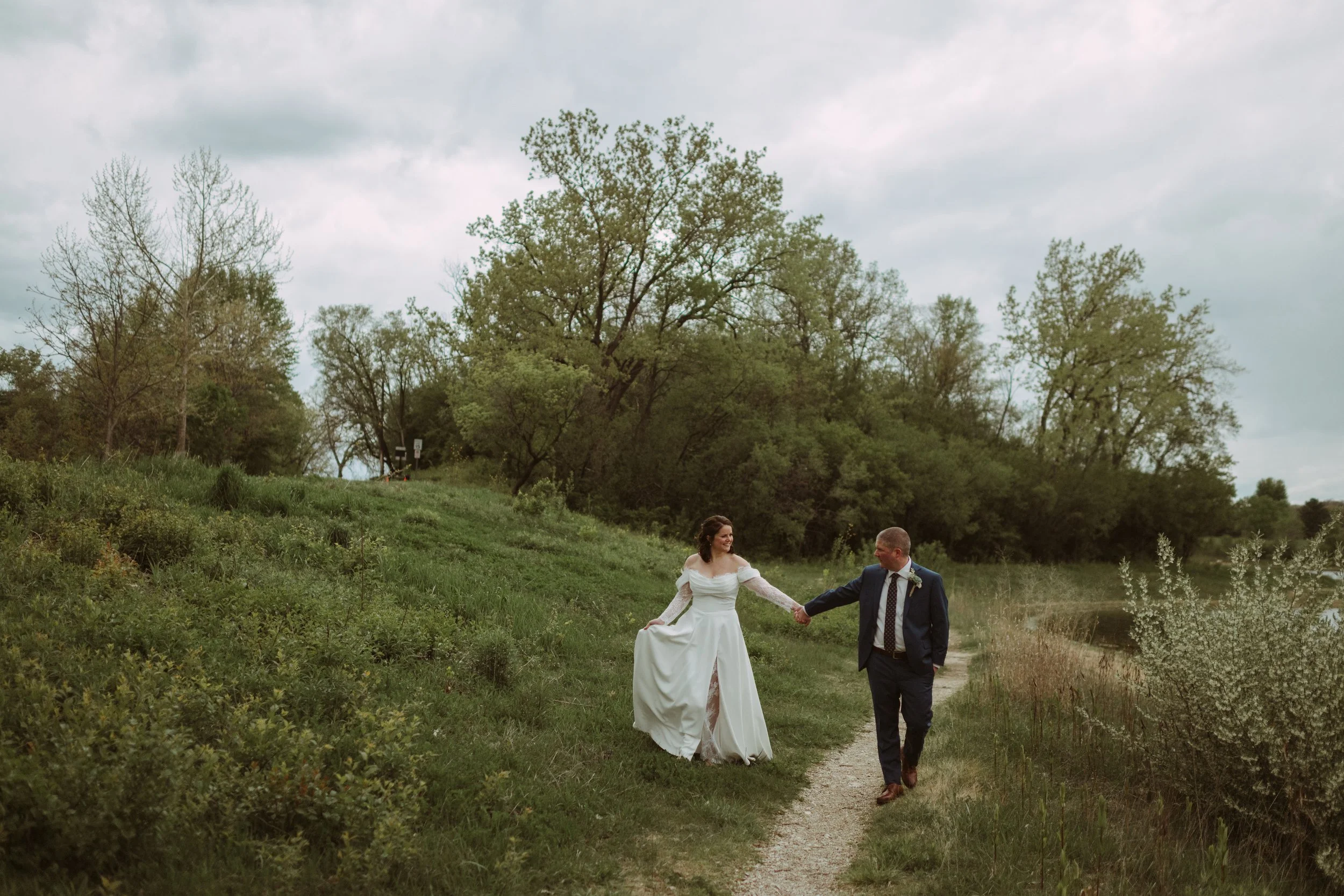 A couple dressed in wedding attire walking hand in hand along a path through a green landscape with trees and a cloudy sky overhead.