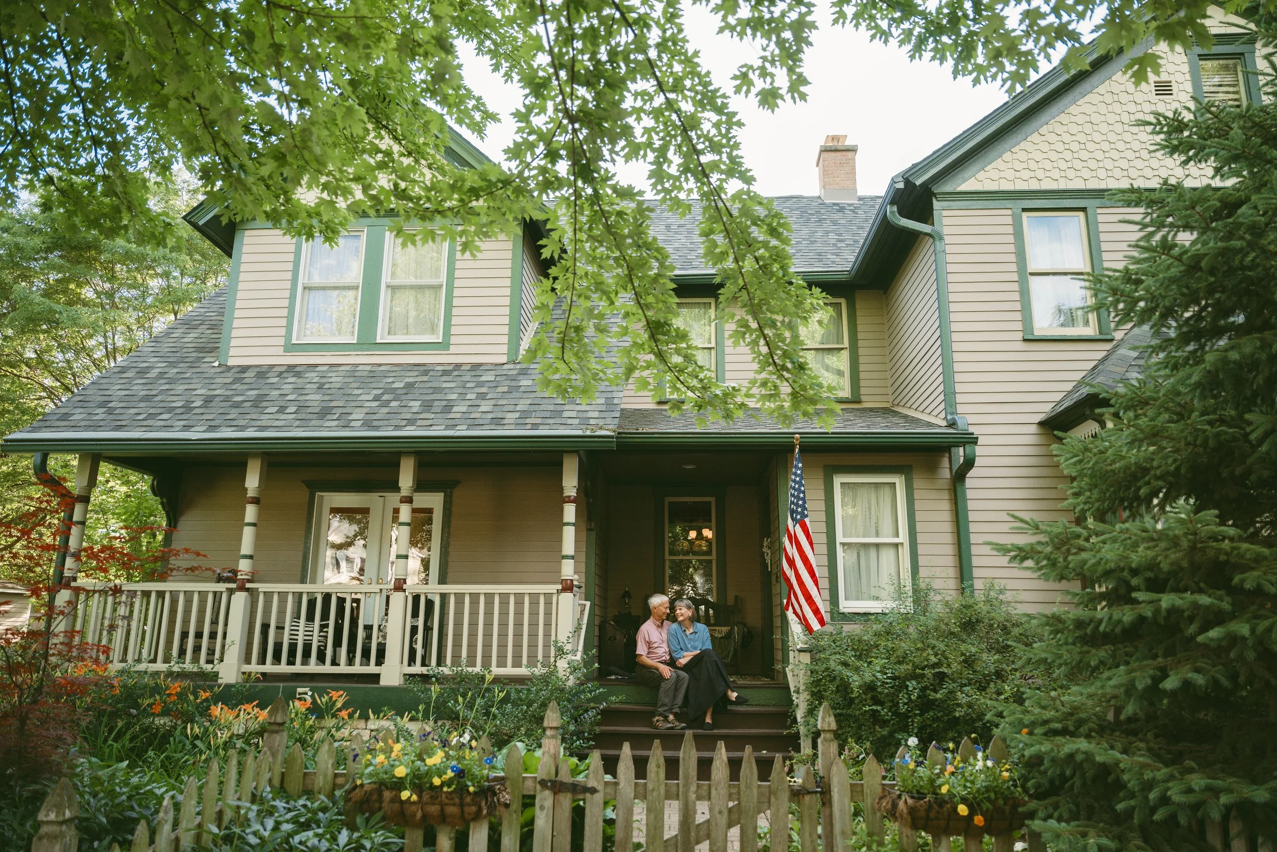 Two elderly women sitting on a porch swing of a house, surrounded by a garden with colorful flowers, with trees and a house in the background.