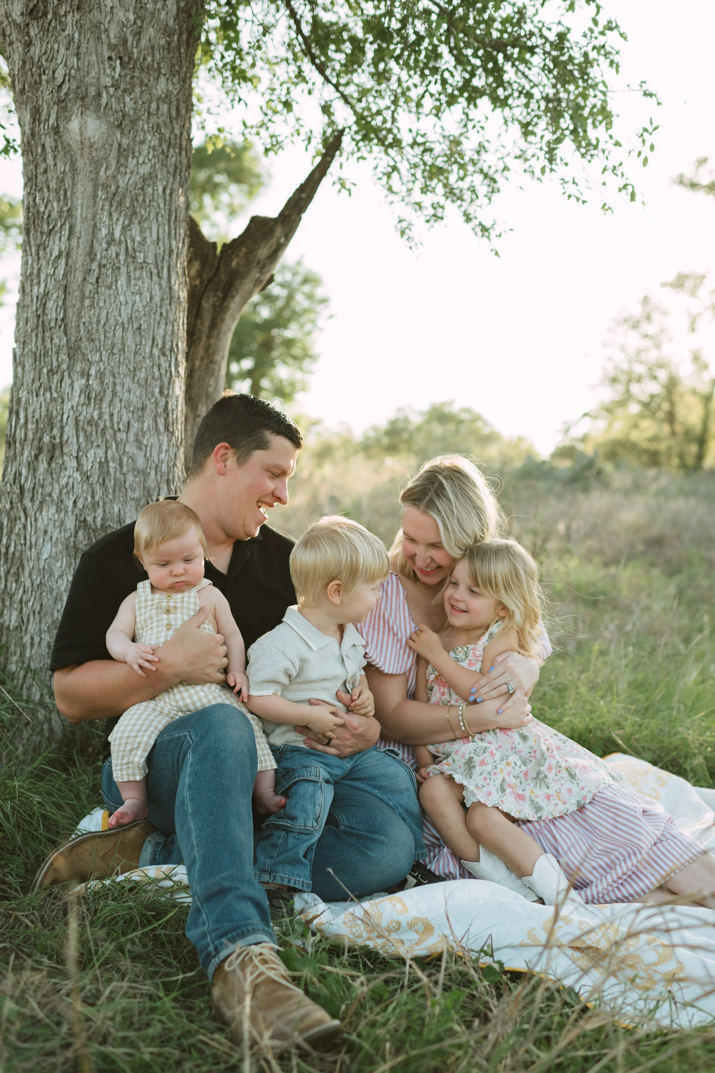 A family with young children sitting on a blanket under a large tree outdoors, smiling and enjoying each other's company.