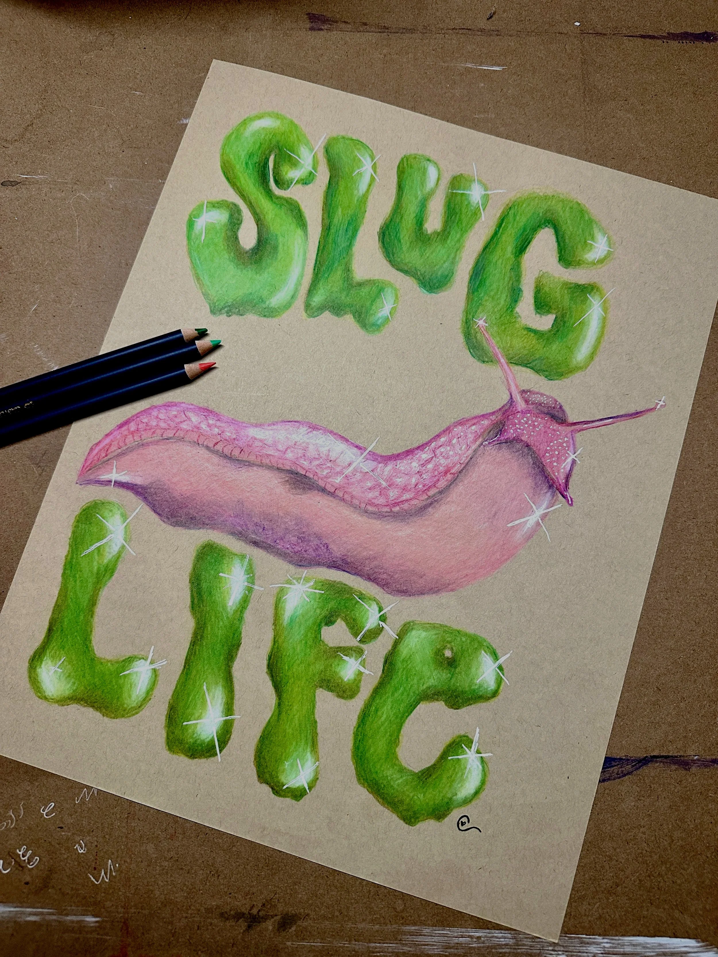 A colorful drawing of the words 'SLUG LIFE' with the letters forming a slime-like texture in green, and a pink slug with a pink shell below the words. The drawing is on beige paper with three colored pencils (blue, black, red) placed beside it.