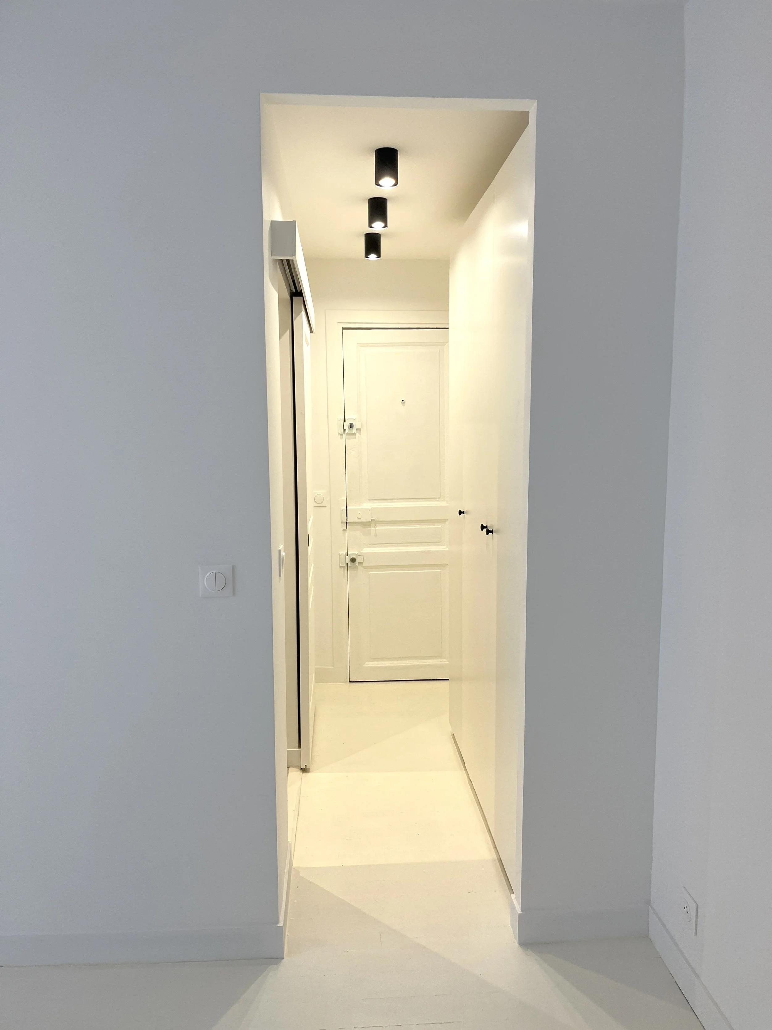 Une vue d'un couloir blanc menant à une porte d'entrée avec un mur blanc et un plafond avec trois lampes encastrées.