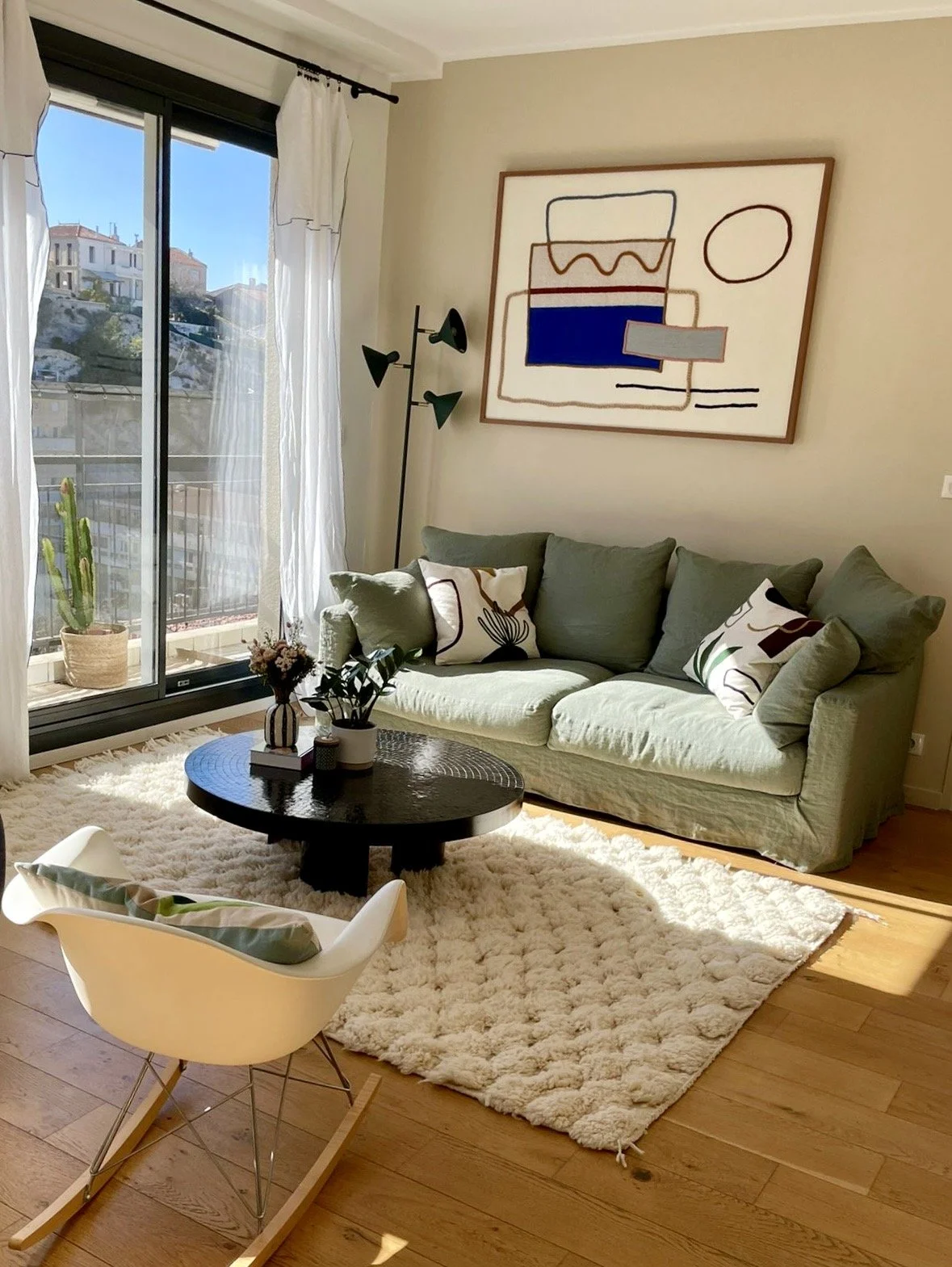 Salon lumineux avec canapé vert, tapis blanc, table basse noire, coussins décoratifs, chaise blanche en bois, grand tableau abstrait sur le mur, grande fenêtre avec rideaux, plante en pot, lampadaire