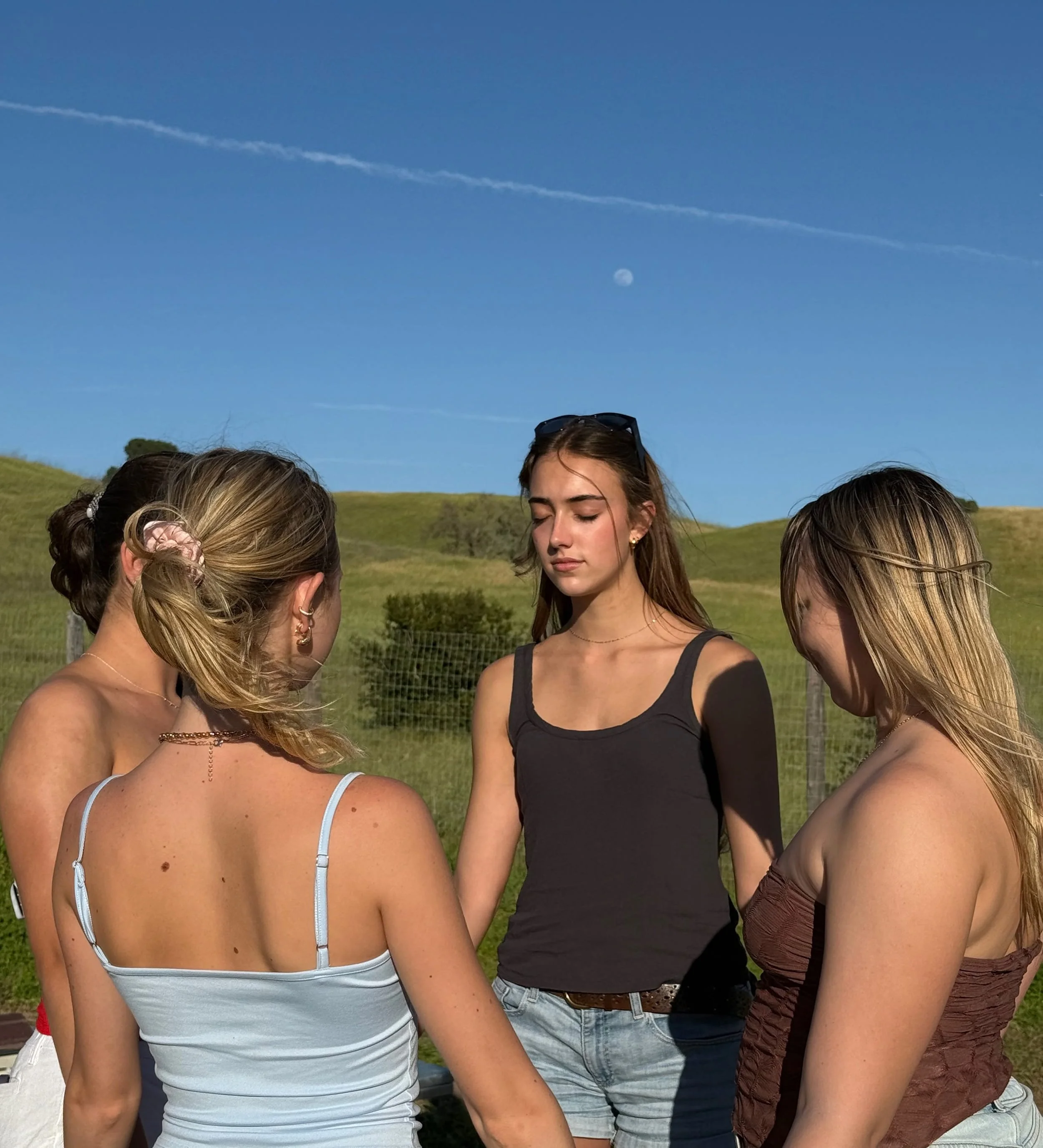 Five young women standing outdoors in a grassy field, with their eyes closed and heads bowed, during daytime with clear blue sky and a visible moon in the background.