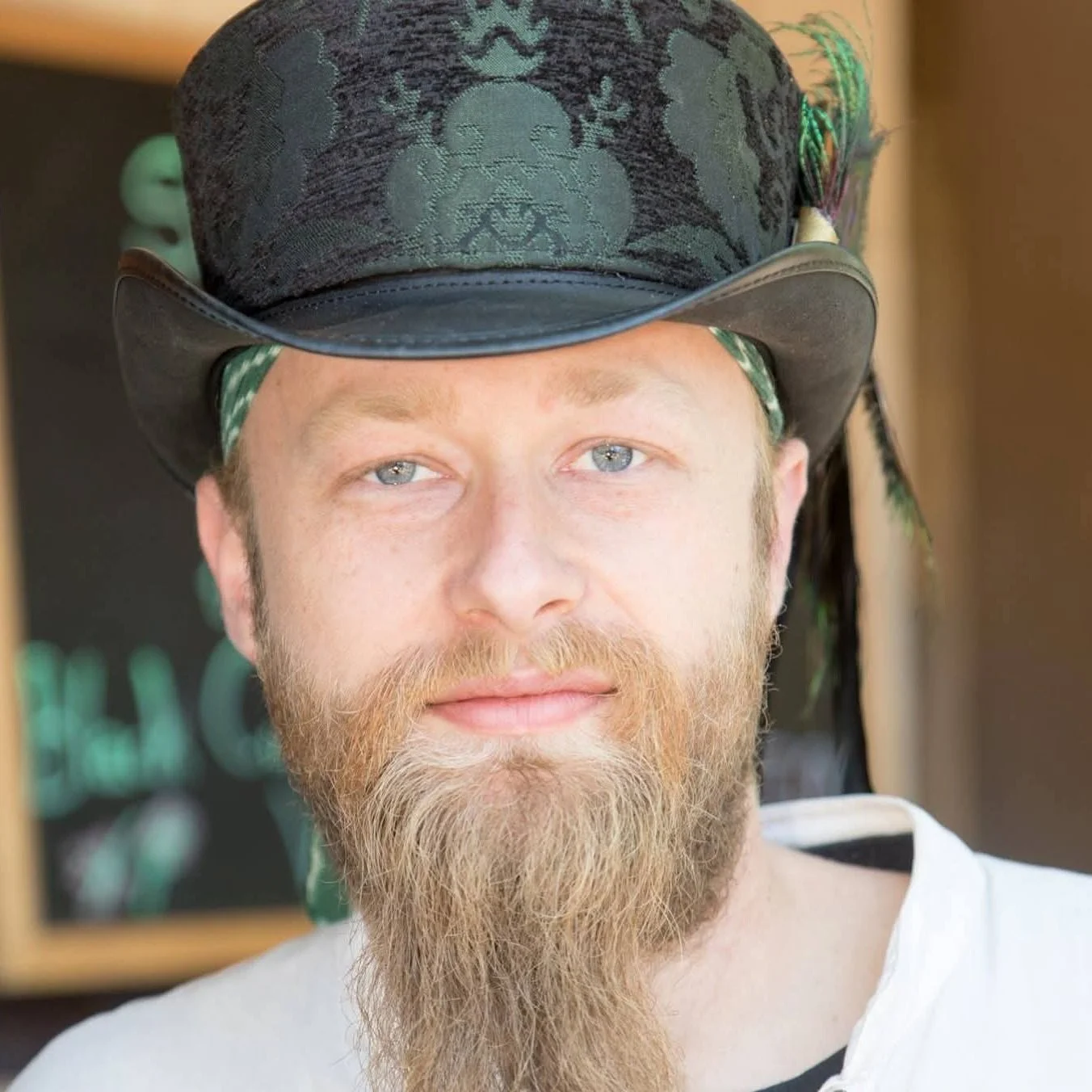 A man with a red beard and blue eyes wearing a black cowboy hat with green embroidery and a white shirt.