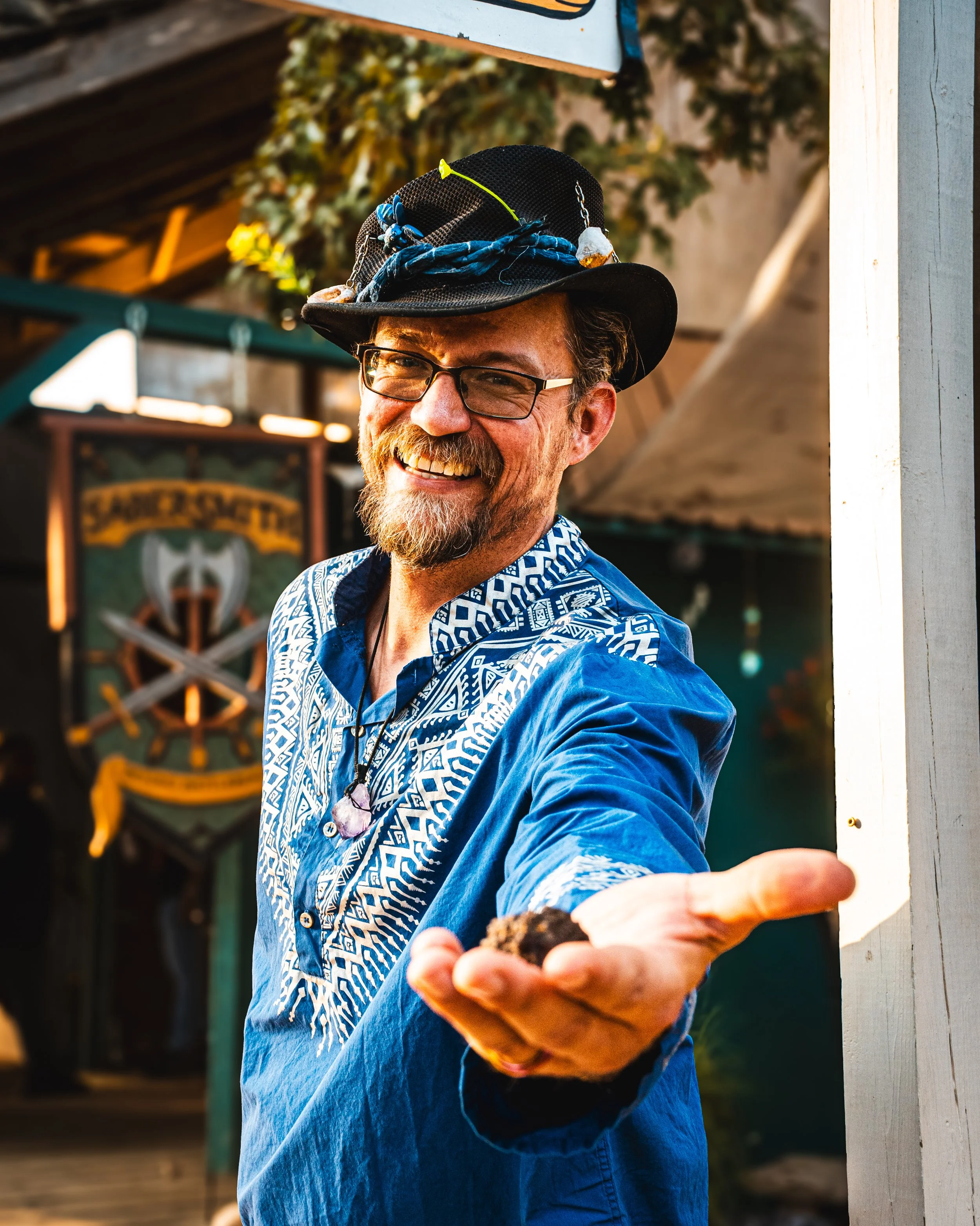 A smiling man in a blue patterned shirt and black hat with decorations, offering a rock to the camera, standing outdoors near a white wall with a sign and a banner in the background.