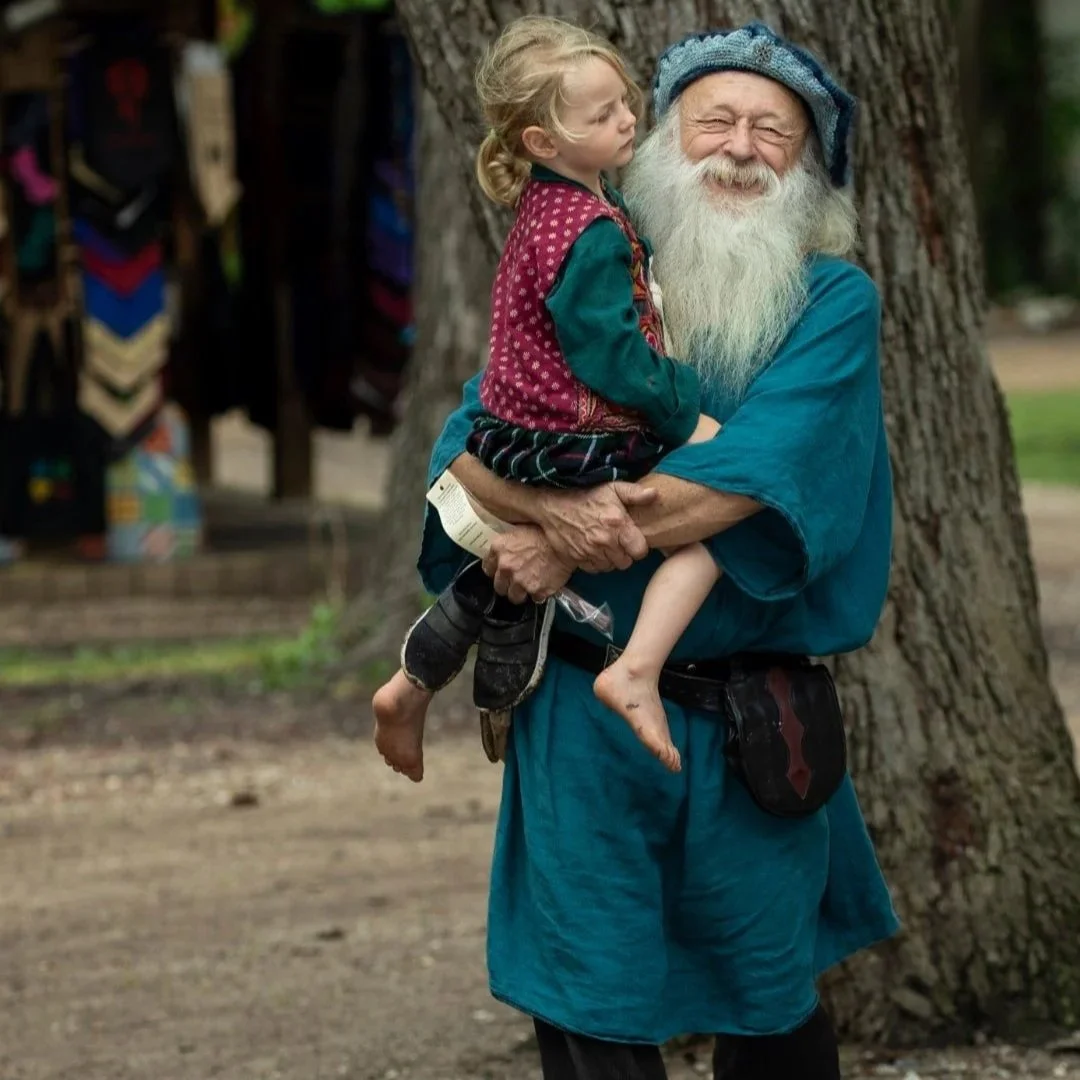 An elderly man with a long white beard wearing a blue tunic and a blue hat, smiling and holding a young girl with blonde hair, who is sitting on his arm, in front of a large tree.