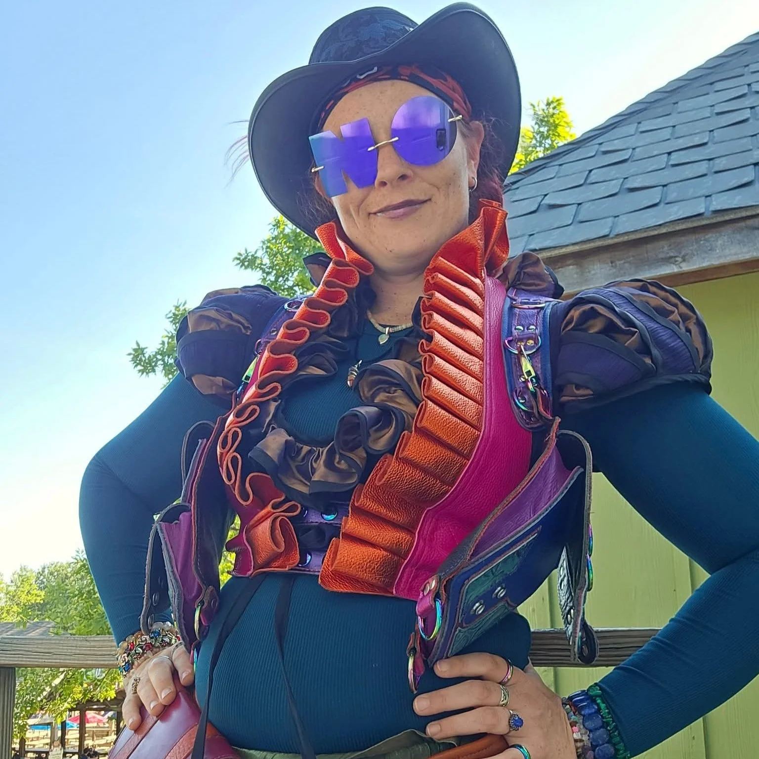 Woman outdoors wearing large purple sunglasses, a black hat, and a colorful ruffled vest, with a blue landscape behind her. The woman's glasses are in the shape of the word NO.
