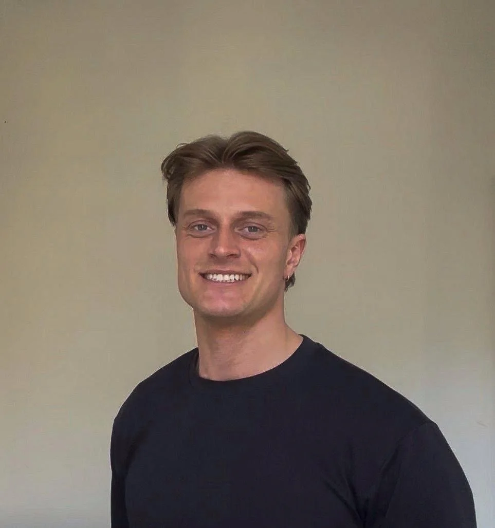 Portret van een lachende jonge man met bruin haar, gekleed in een zwart T-shirt, staand voor een effen lichtgele achtergrond.