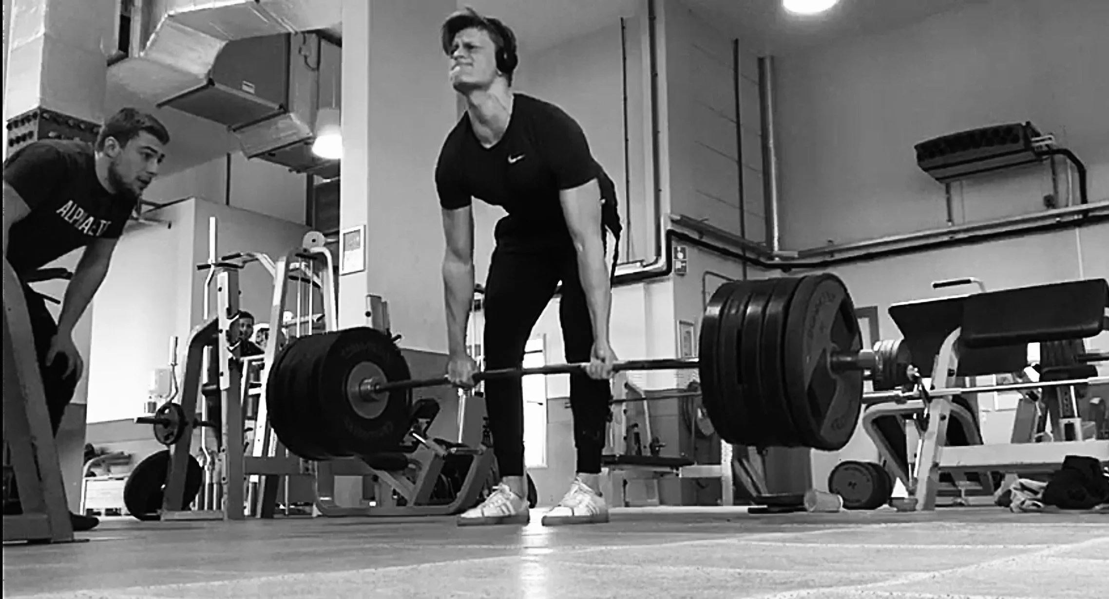 Man tilt deadlift met zware gewichten in een sportschool, omringd door apparatuur. Een man kijkt geconcentreerd terwijl hij de oefening uitvoert, met twee mensen die hem begeleiden of toekijken. In Scheveningen, Den Haag.