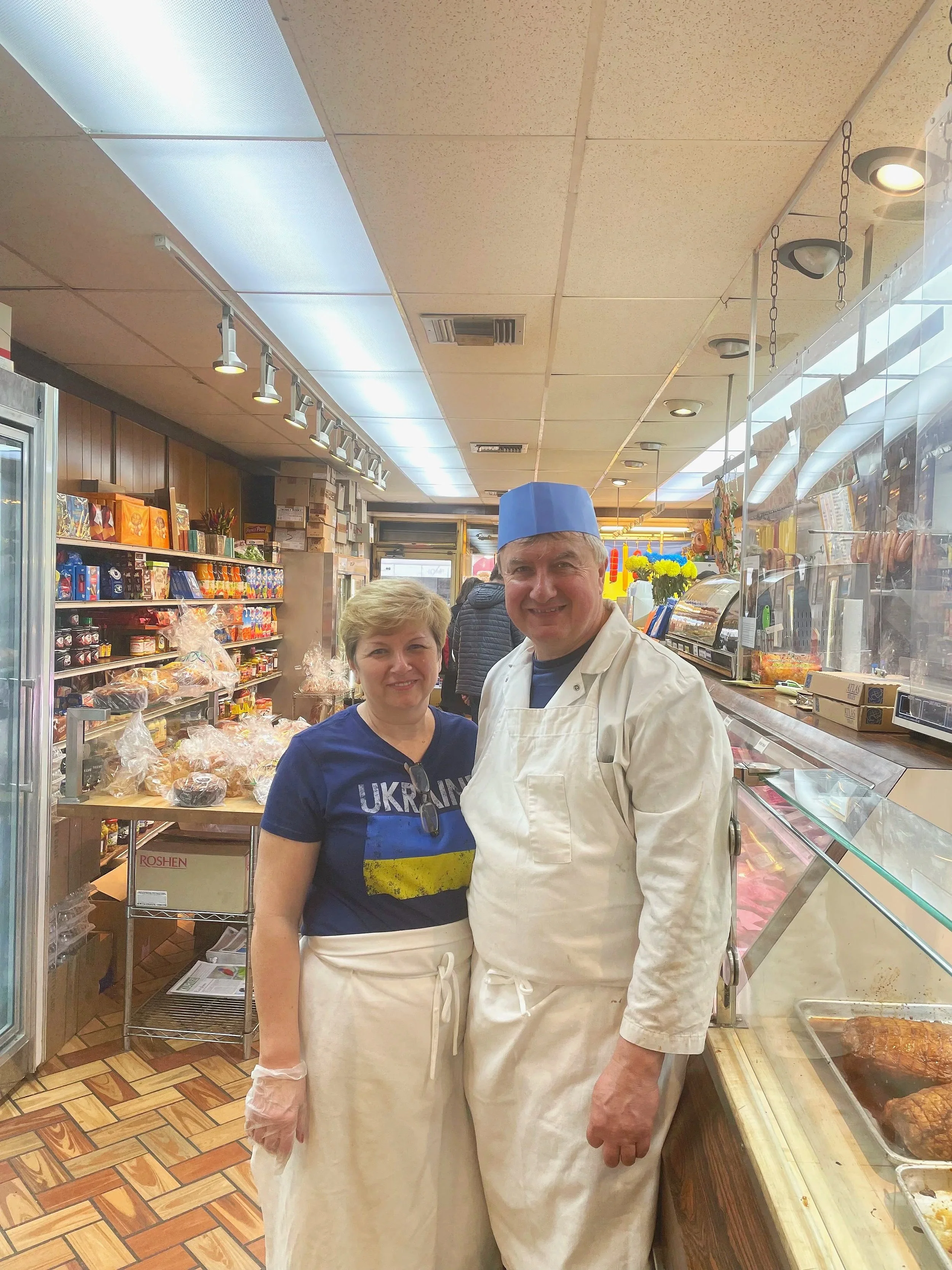 Two people, a woman and a man, smile at the camera inside a grocery store. The woman wears a blue Ukraine shirt and plastic gloves, while the man wears a blue cap, white apron, and dress shirt. Store shelves with baked goods, candies, and flowers are visible behind them.