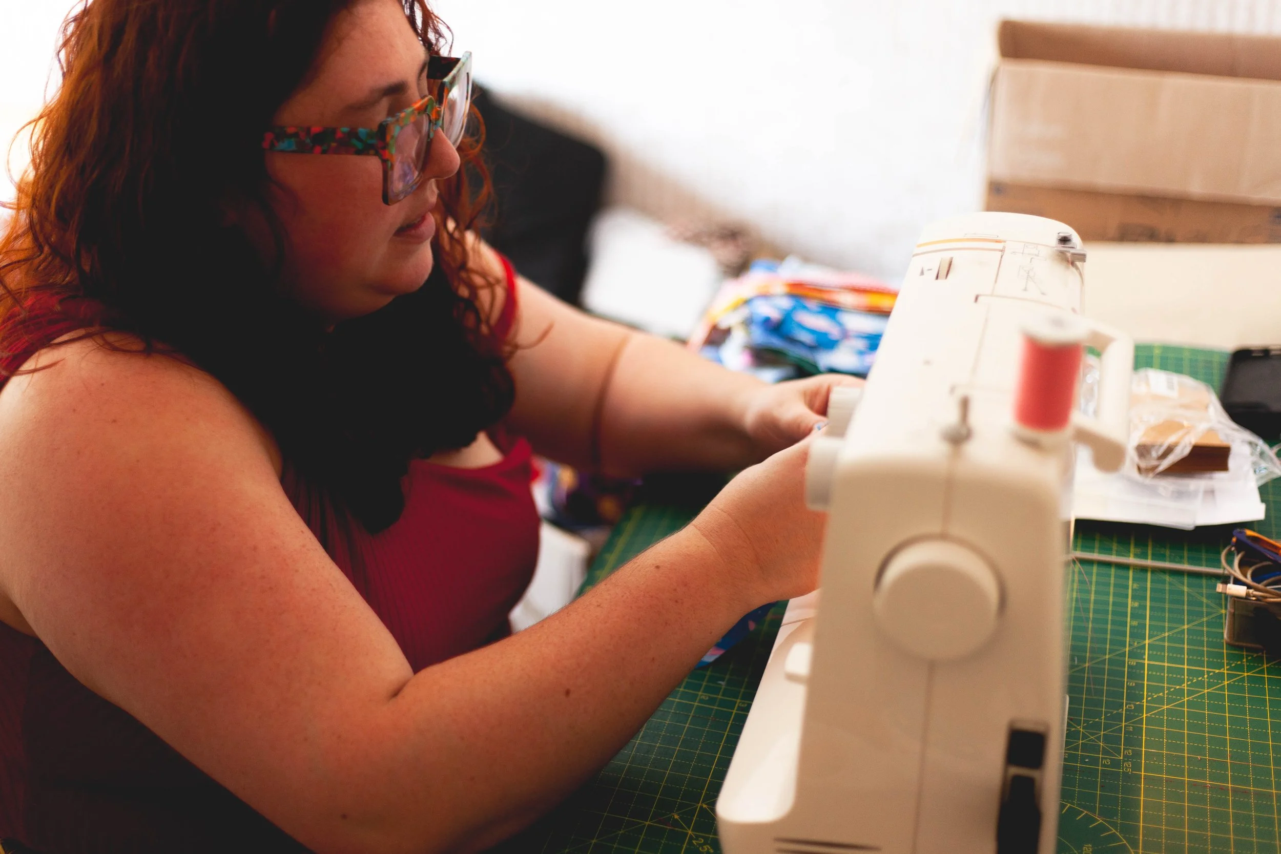 A woman with red hair, wearing glasses and a red sleeveless top, is sewing on a white sewing machine at a green cutting mat table surrounded by fabrics and sewing supplies.