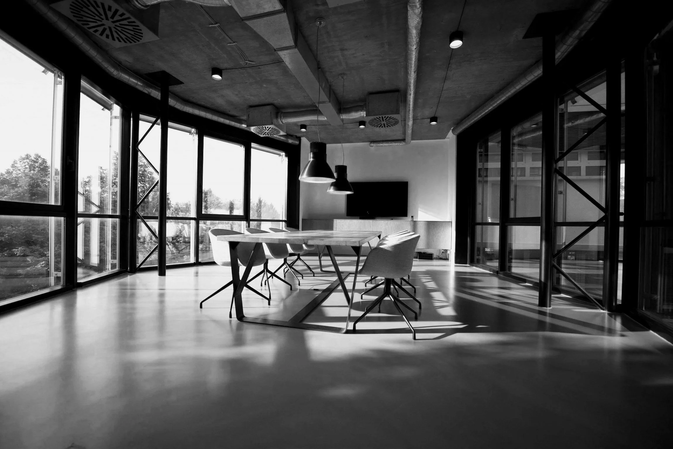 Interior of a modern office with large windows, concrete ceiling with ductwork, hanging lights, a wall-mounted TV, and a wooden table with chairs.