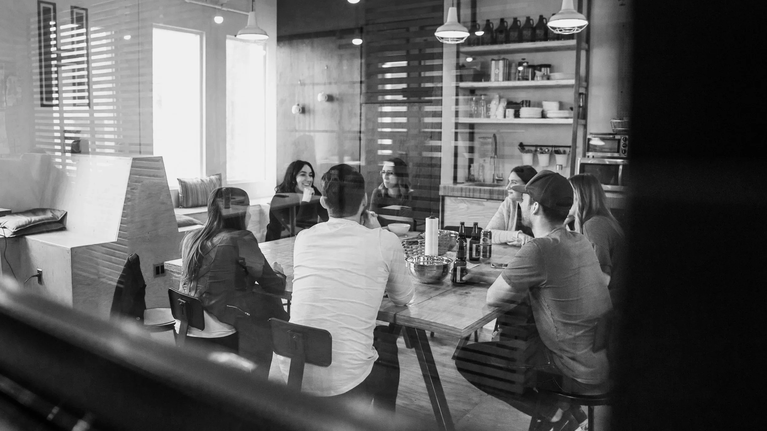 A group of people sitting around a wooden table in a cozy cafe or restaurant, enjoying drinks and conversation. The scene is viewed through a glass window, with the cafe's interior featuring shelves with dishes, bottles, and hanging lights.