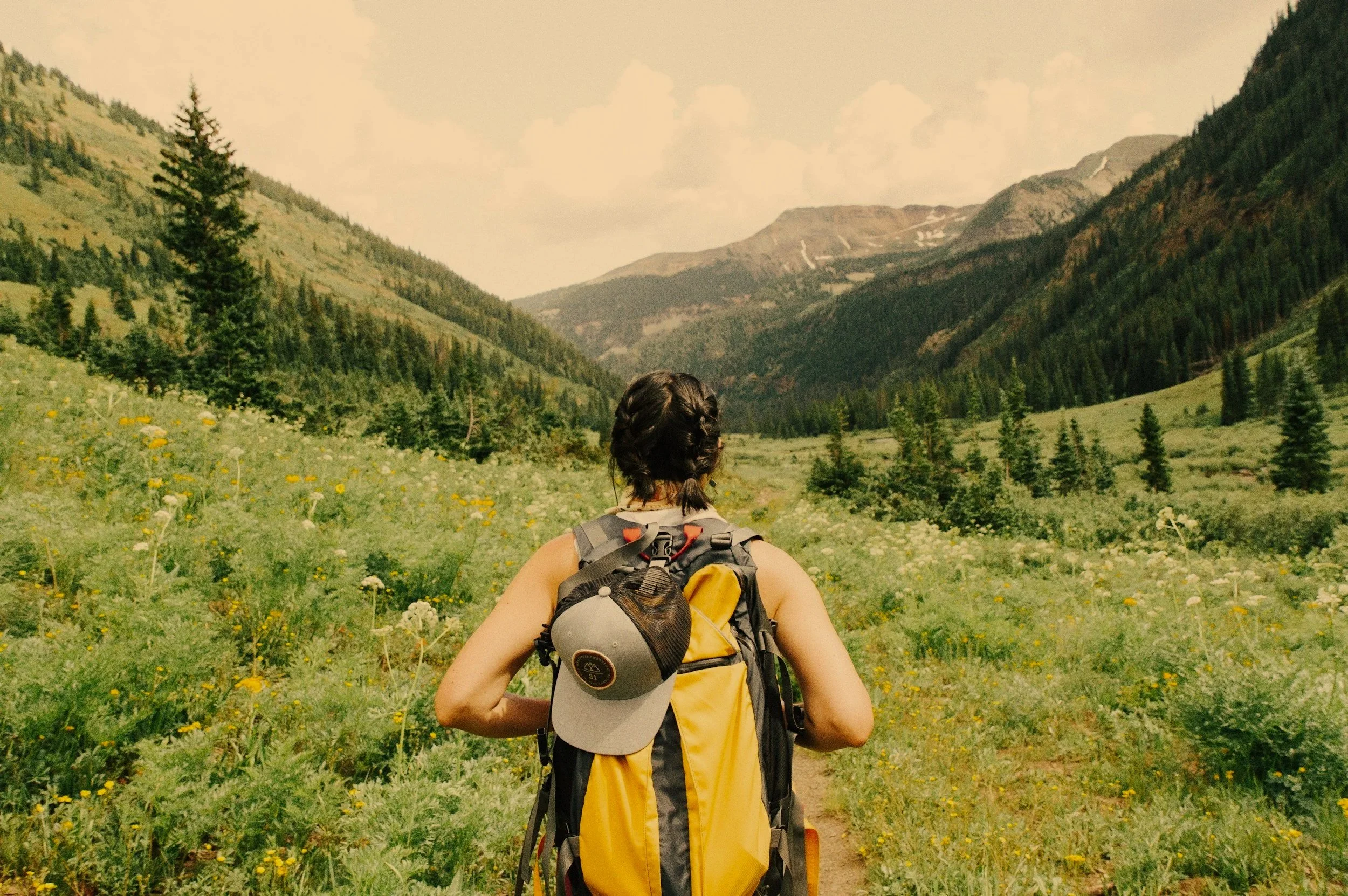 A woman with braided dark hair hiking on a trail in a lush green valley surrounded by mountains, carrying a yellow and black backpack with a gray cap hanging on it, during daytime with a partly cloudy sky.