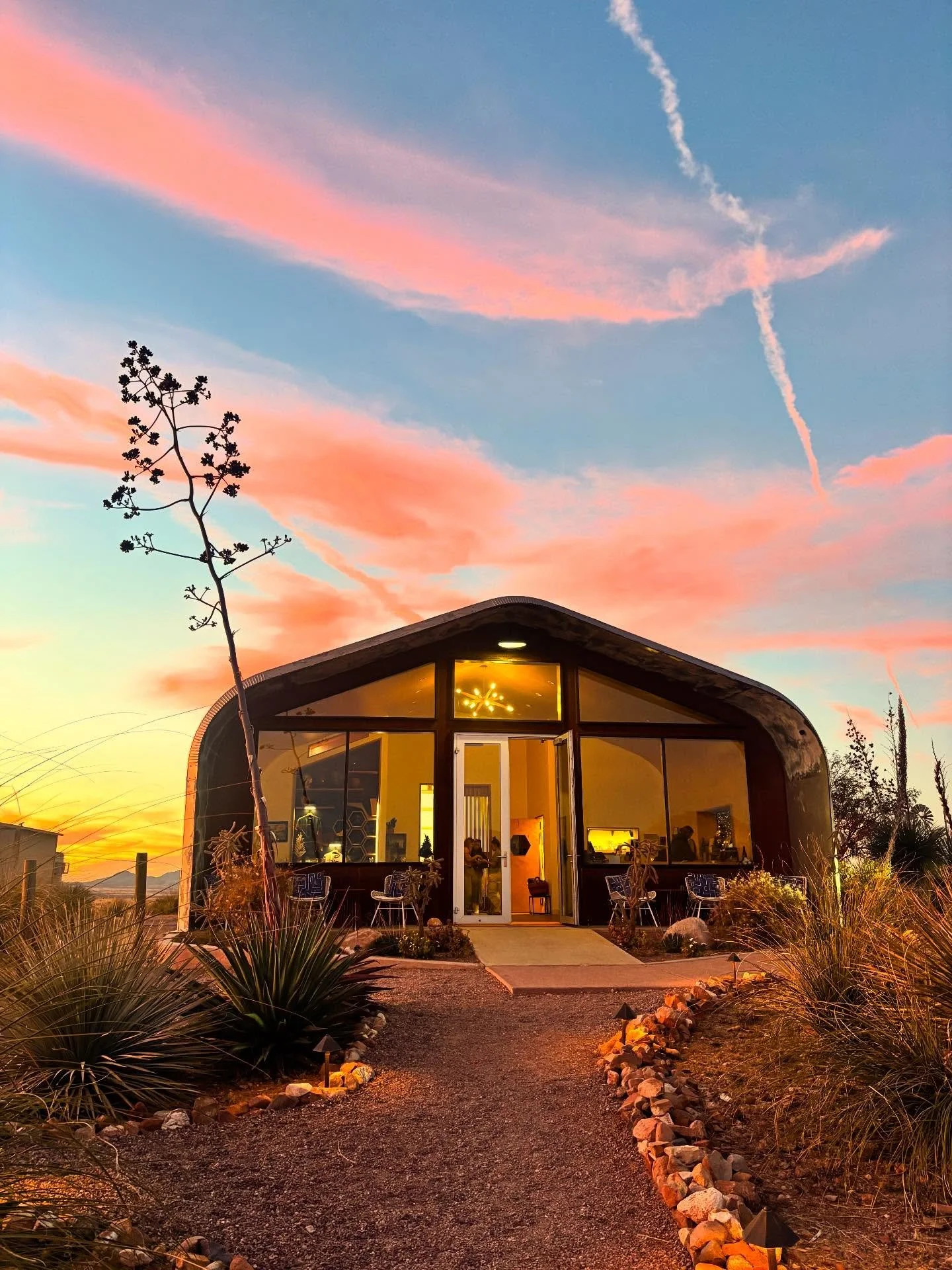 Rune A modern house with large glass windows, illuminated from inside, situated in a desert-like landscape at sunset with colorful pink and orange clouds in the sky.