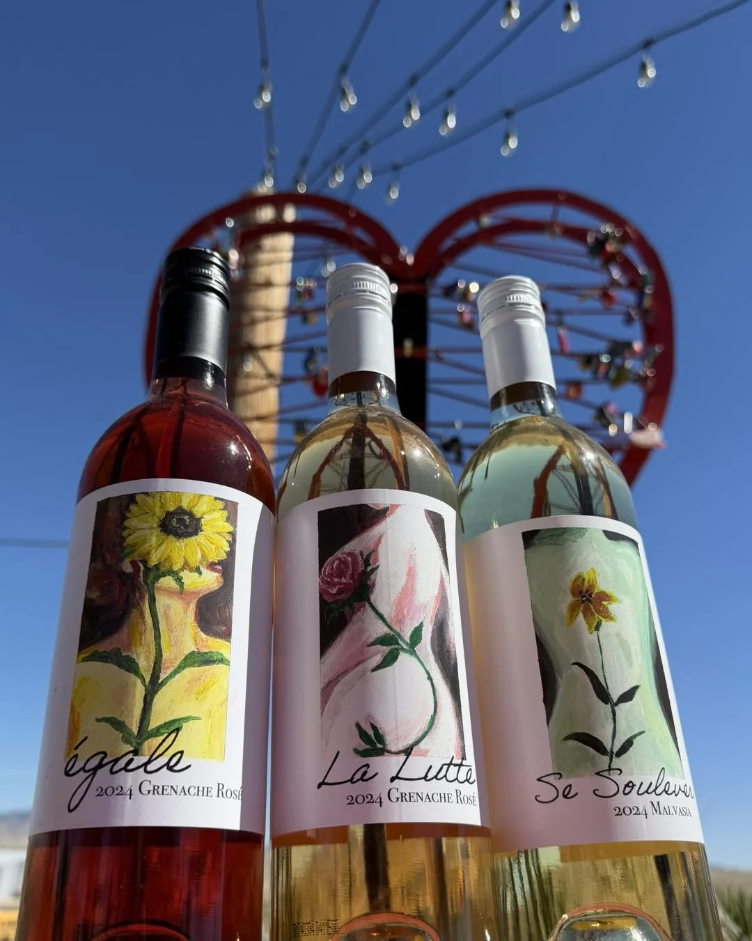 Three bottles of rosé wine with artistic labels, set against a clear blue sky and a red Ferris wheel in the background.