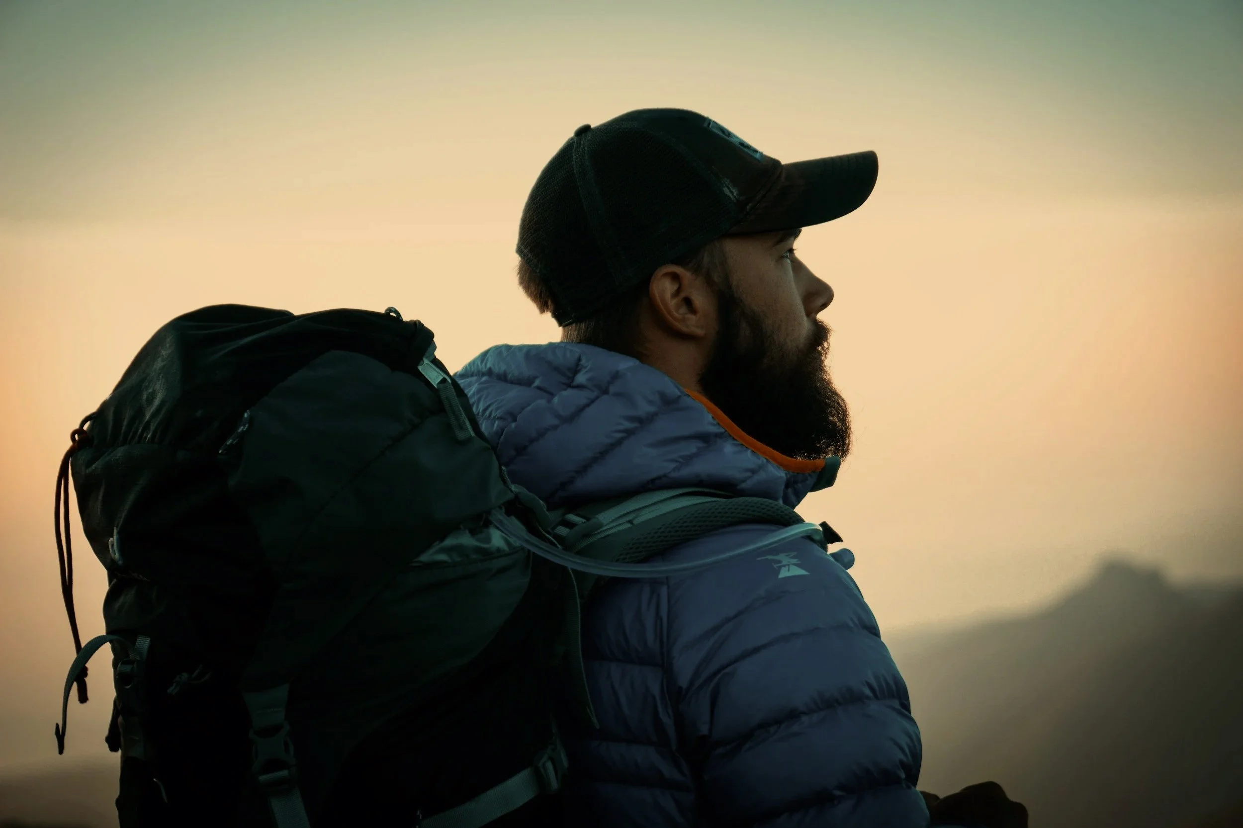 A man with a beard wearing a baseball cap, blue puffer jacket, and carrying a backpack outdoors during sunset or sunrise.