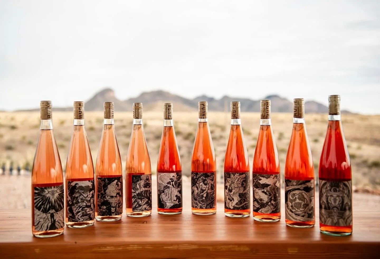 Rune Winery A row of nine bottles of rosé wine with artistic black and white labels, set on a wooden surface outdoors with a mountainous landscape in the background.