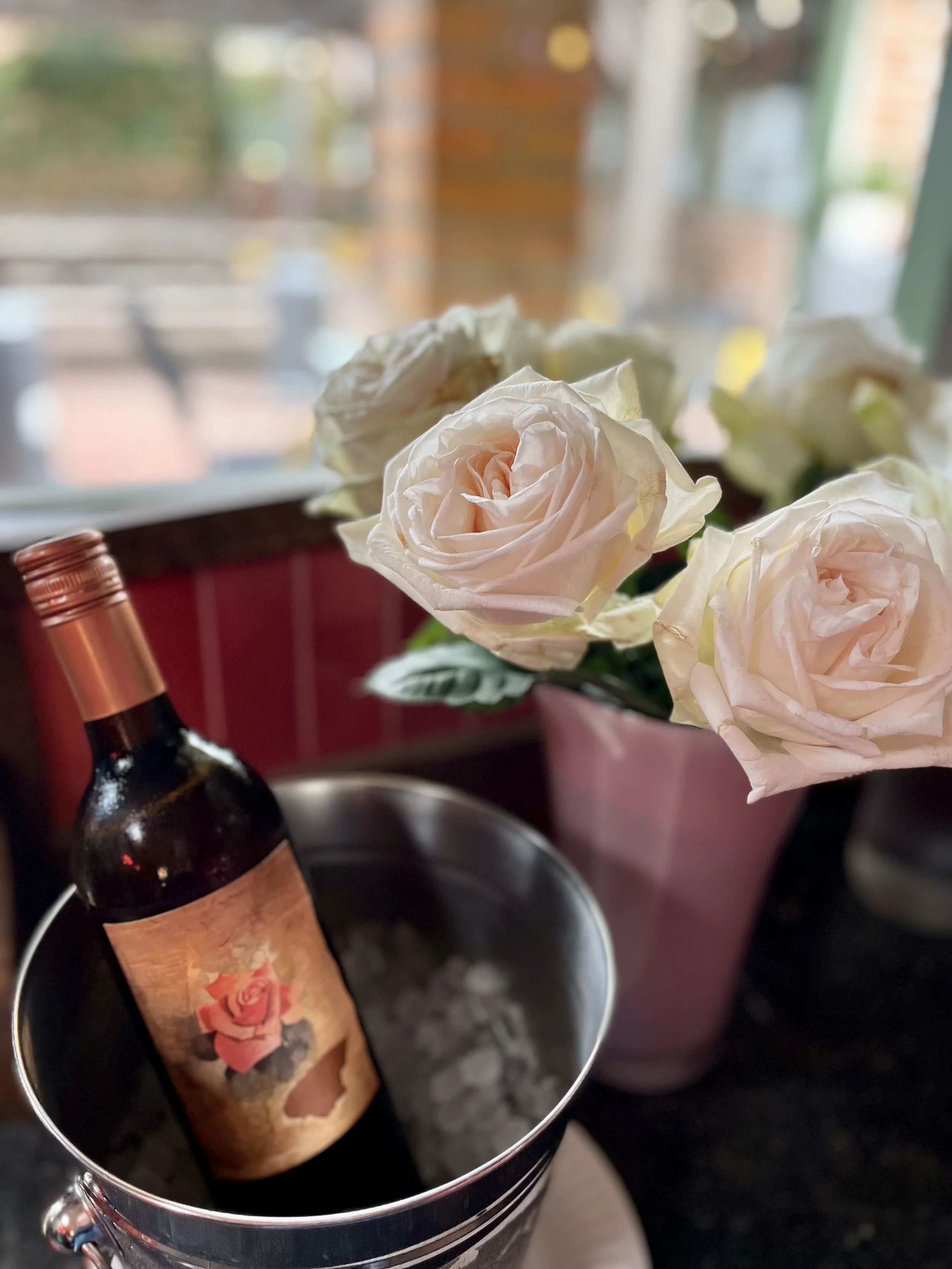 A bottle of red wine with a rose label inside a silver ice bucket, placed next to a pink pot of white roses on a countertop near a window with blurred outdoor scene behind.