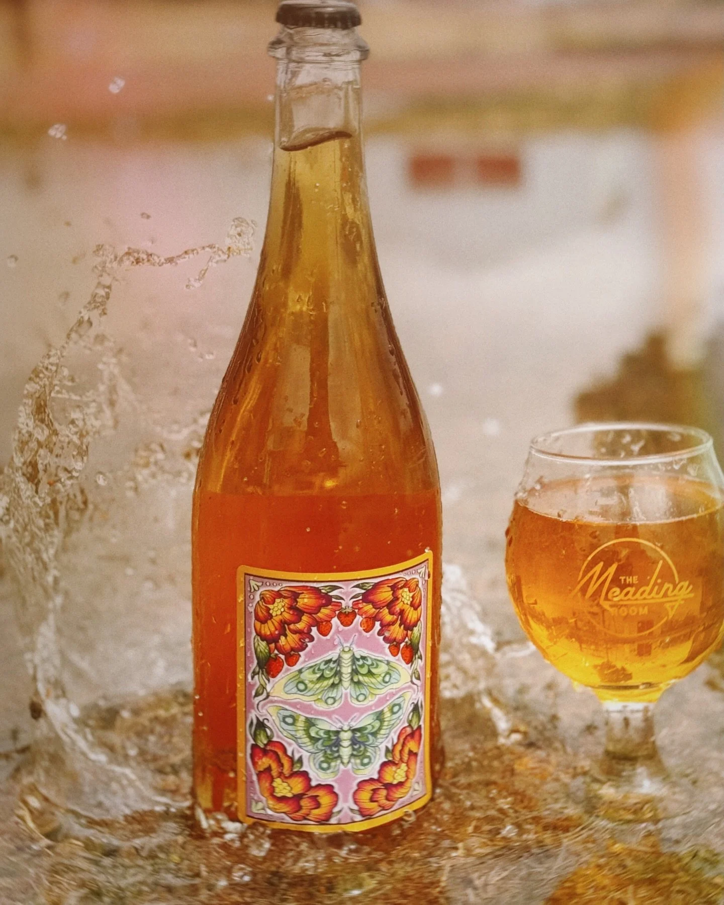A glass and a bottle of orange-colored beverage with a colorful butterfly and floral design label, surrounded by ice with water splashing around.