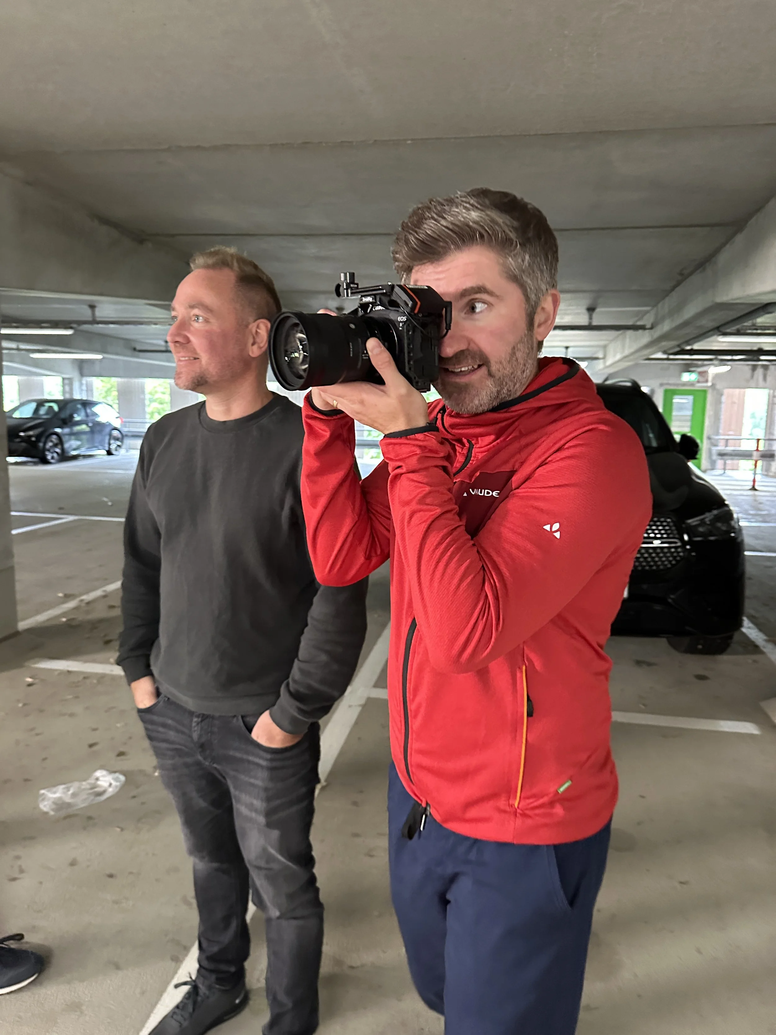 Zwei Männer in einem Parkhaus, einer mit einer Kamera aufblickend, der andere lächelnd und mit Händen in den Taschen.