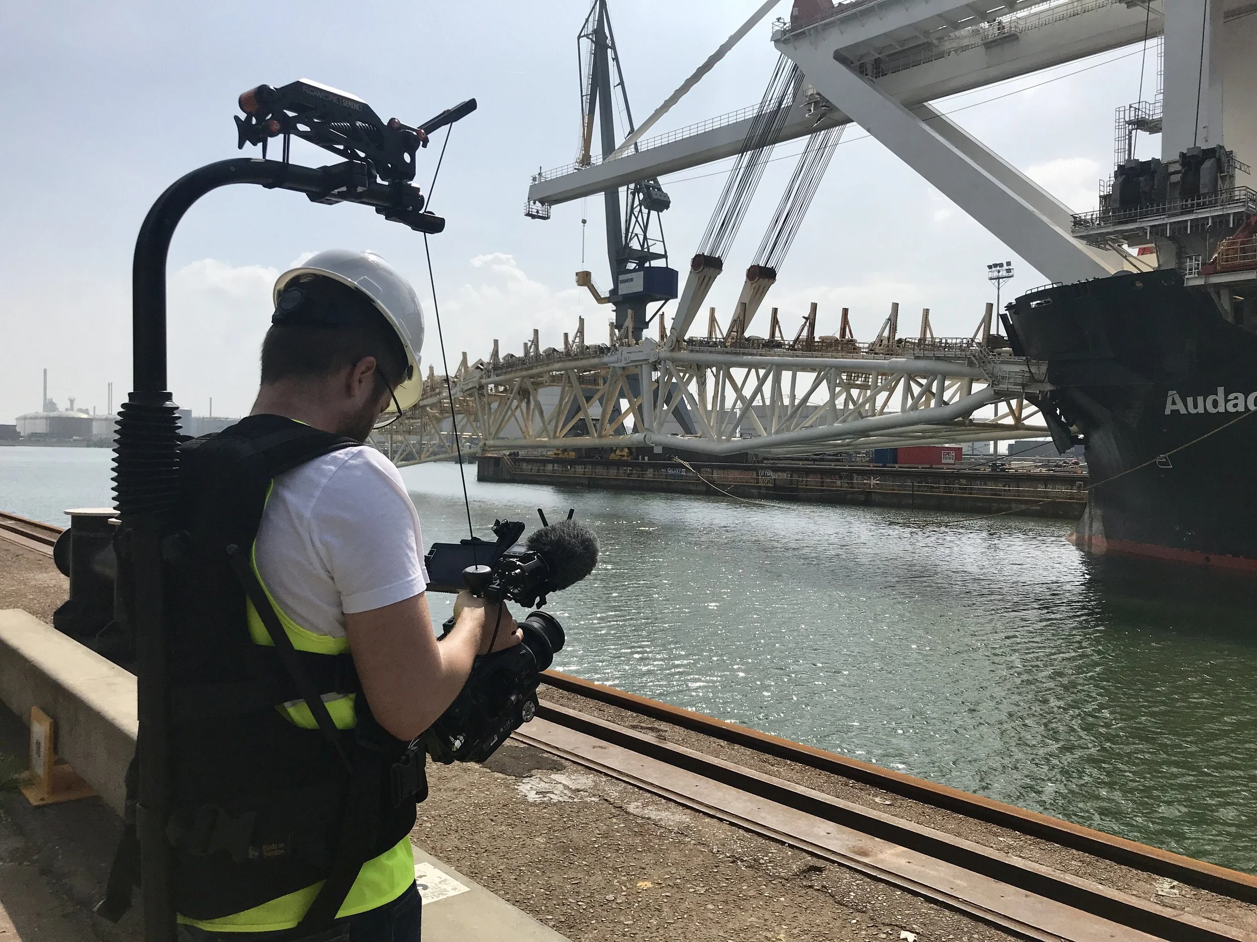 Ein Reporterin filmt mit einer Kamera an einem Hafen, im Hintergrund liegt ein großes Schiff und eine Krananlage.