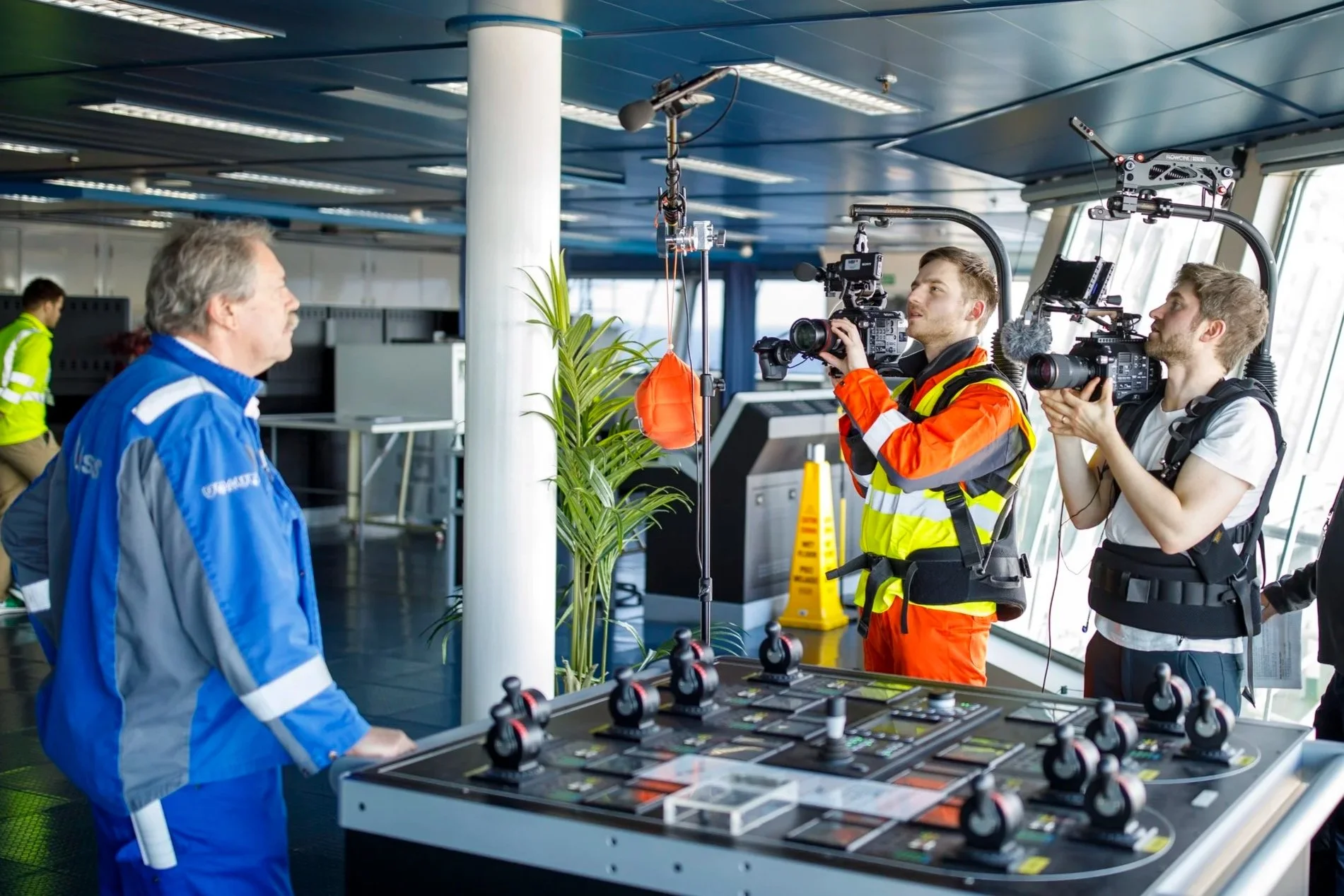 Ein Mann in blauer Jacke wird im Gespräch mit zwei Filmemachern, die Kameras auf ihn richten, in einem modernen, gut beleuchteten Raum mit großen Fenstern und Technik im Hintergrund.