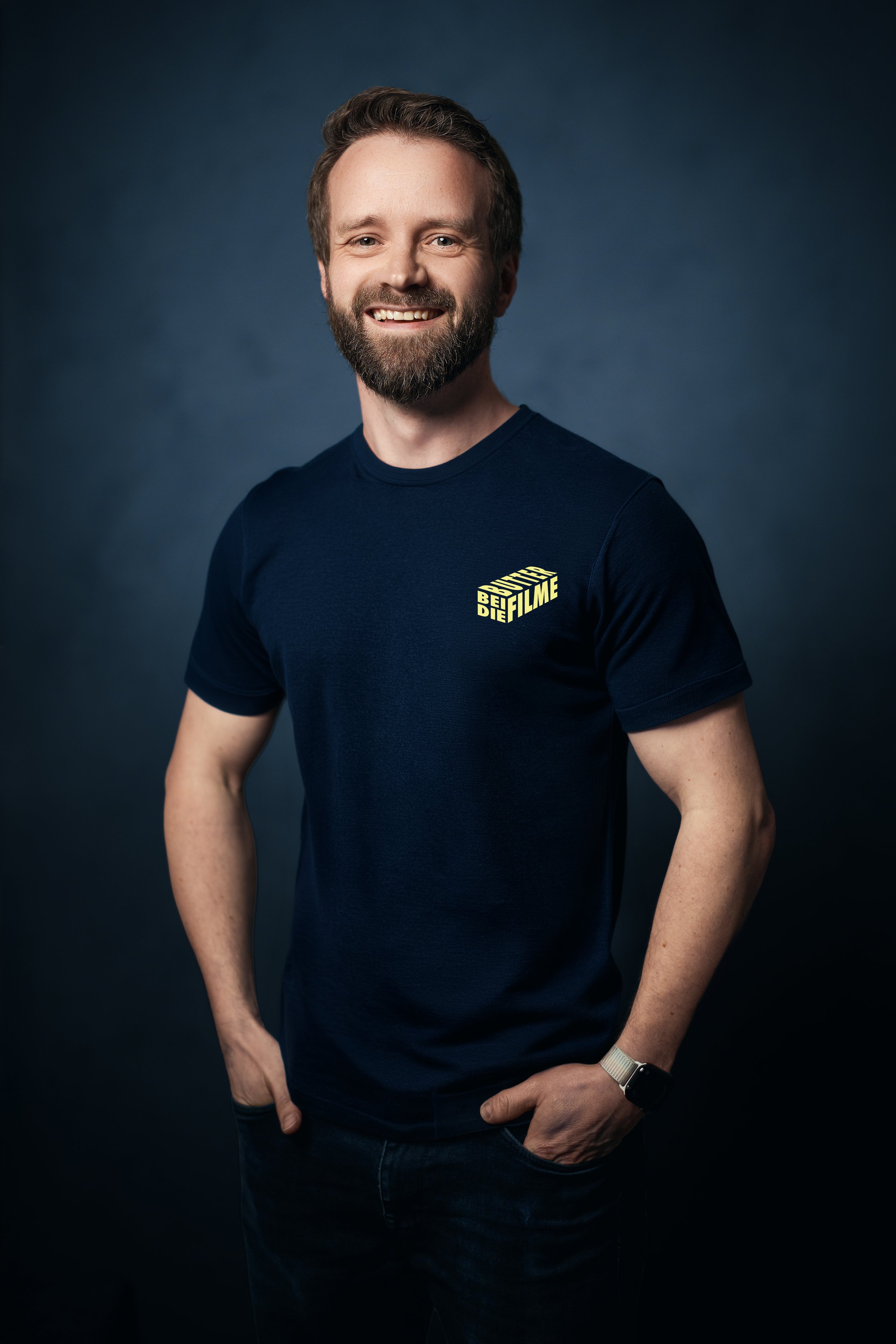 Lächelnder Mann mit braunem Haar und Bart, trägt ein dunkelblaues T-Shirt mit gelbem Logo, steht gegen einen dunklen Hintergrund.