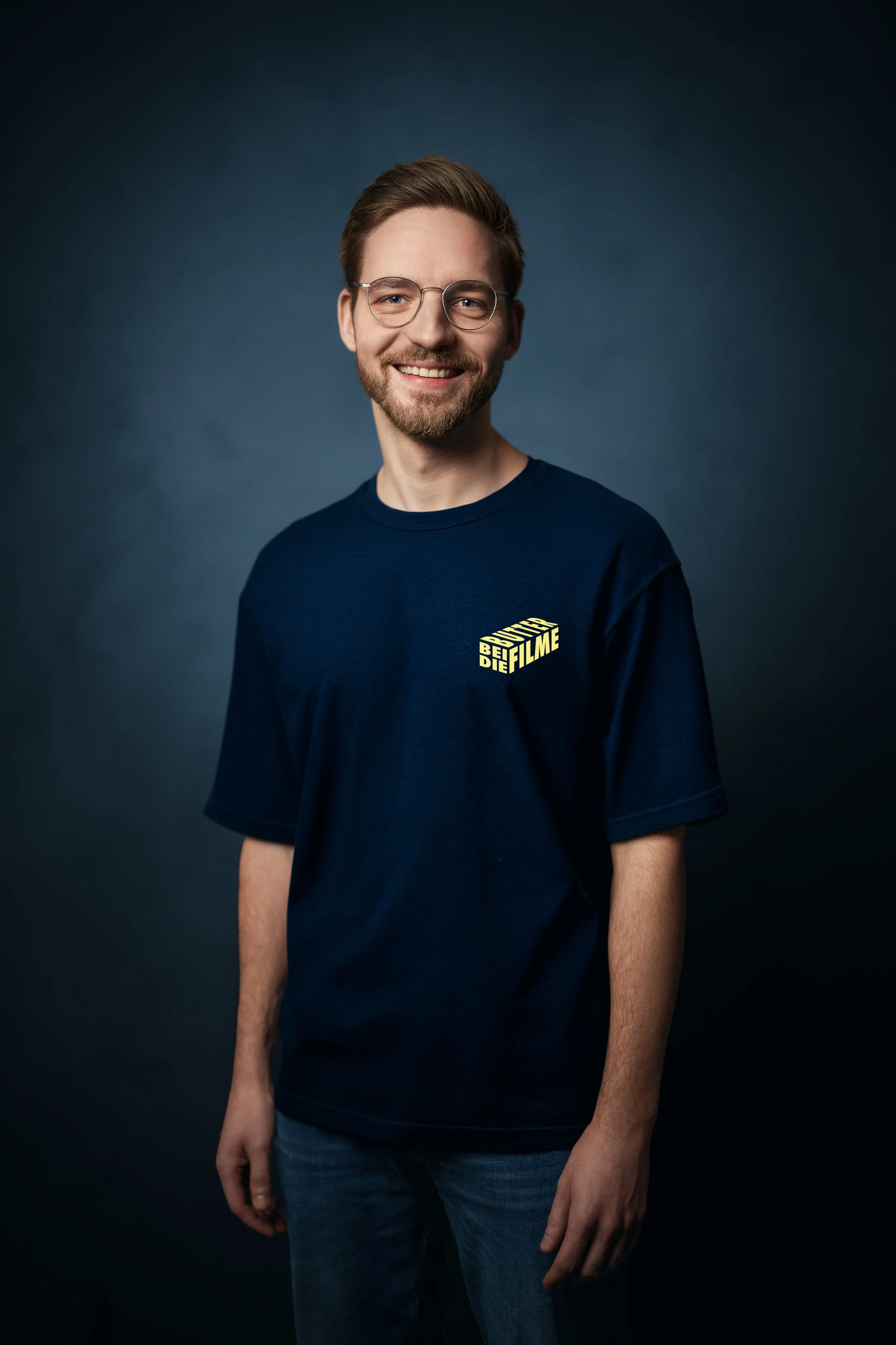 Ein junger Mann mit Brille, schlägt ein Lächeln auf, trägt ein dunkelblaues T-Shirt mit gelbem Aufdruck und steht vor dunklem Hintergrund.