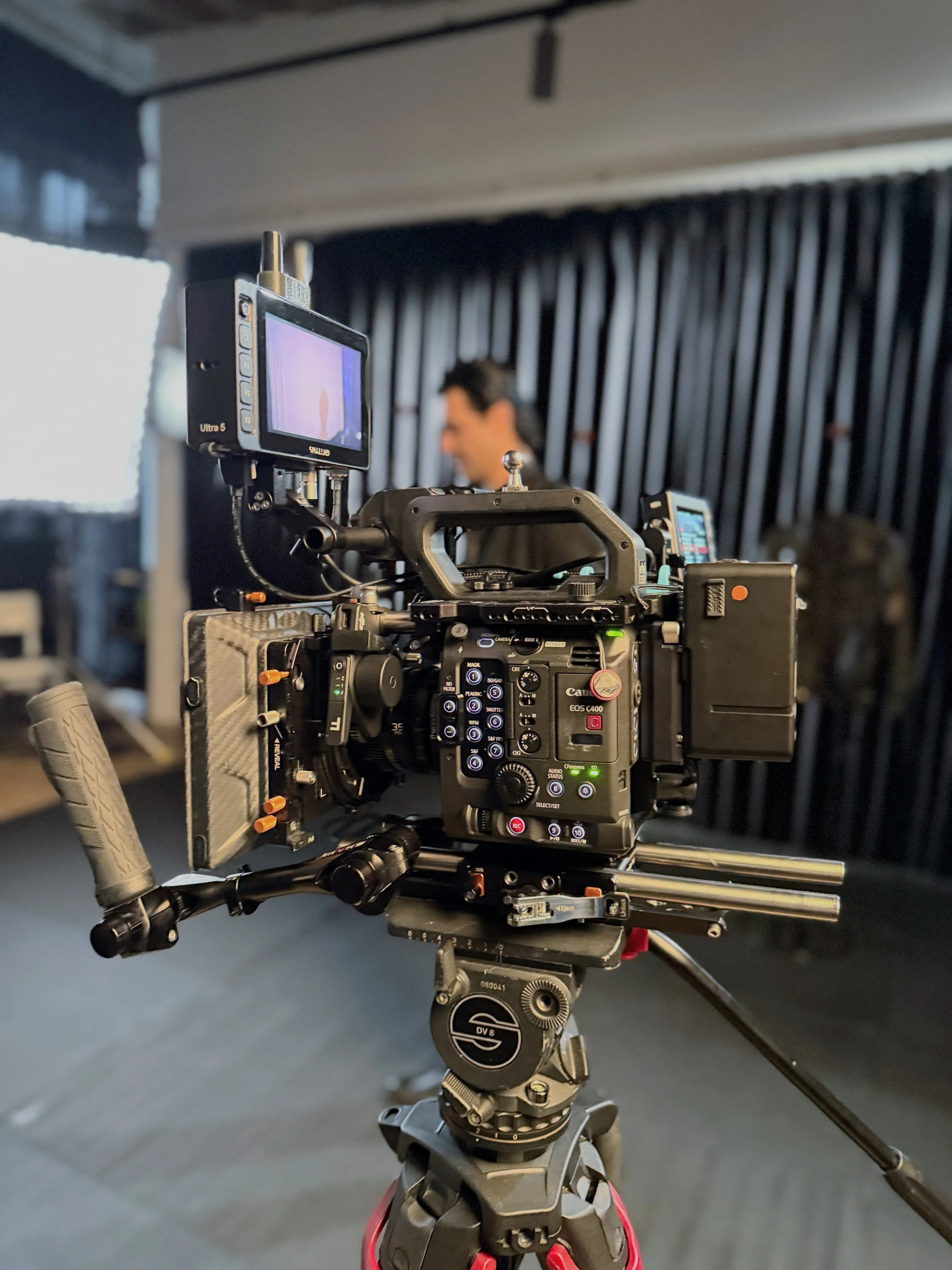 Professionelle Filmkamera auf einem Stativ in einem Studio, im Hintergrund eine Person, kaum erkennbar, mit schwarzem Hintergrund und Lichtreflexen.