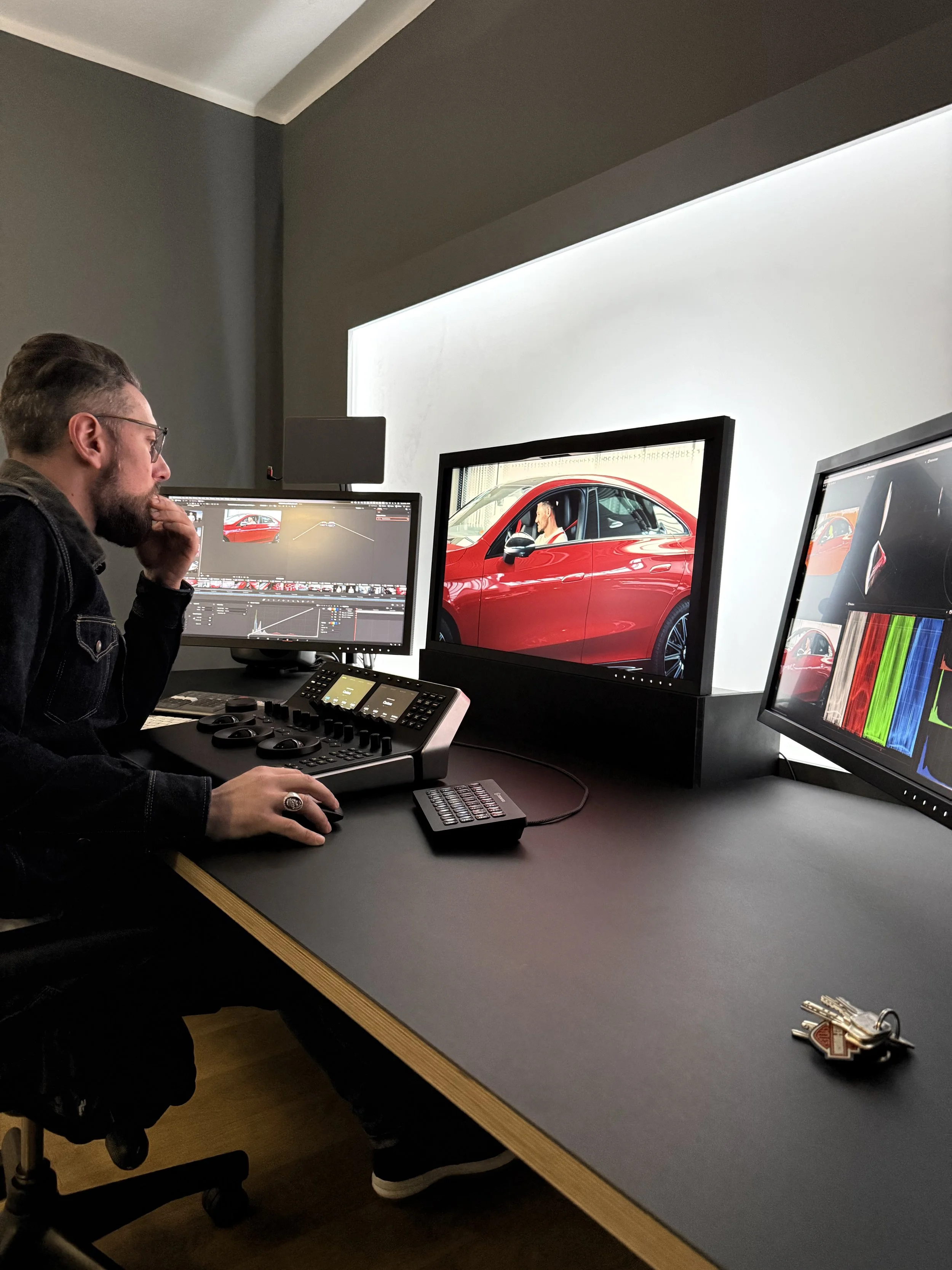 Ein Mann arbeitet an einem Schnittstellencomputer in einem dunklen Raum, während er einen rot lackierten Tesla im Hintergrund auf einem Monitor betrachtet.