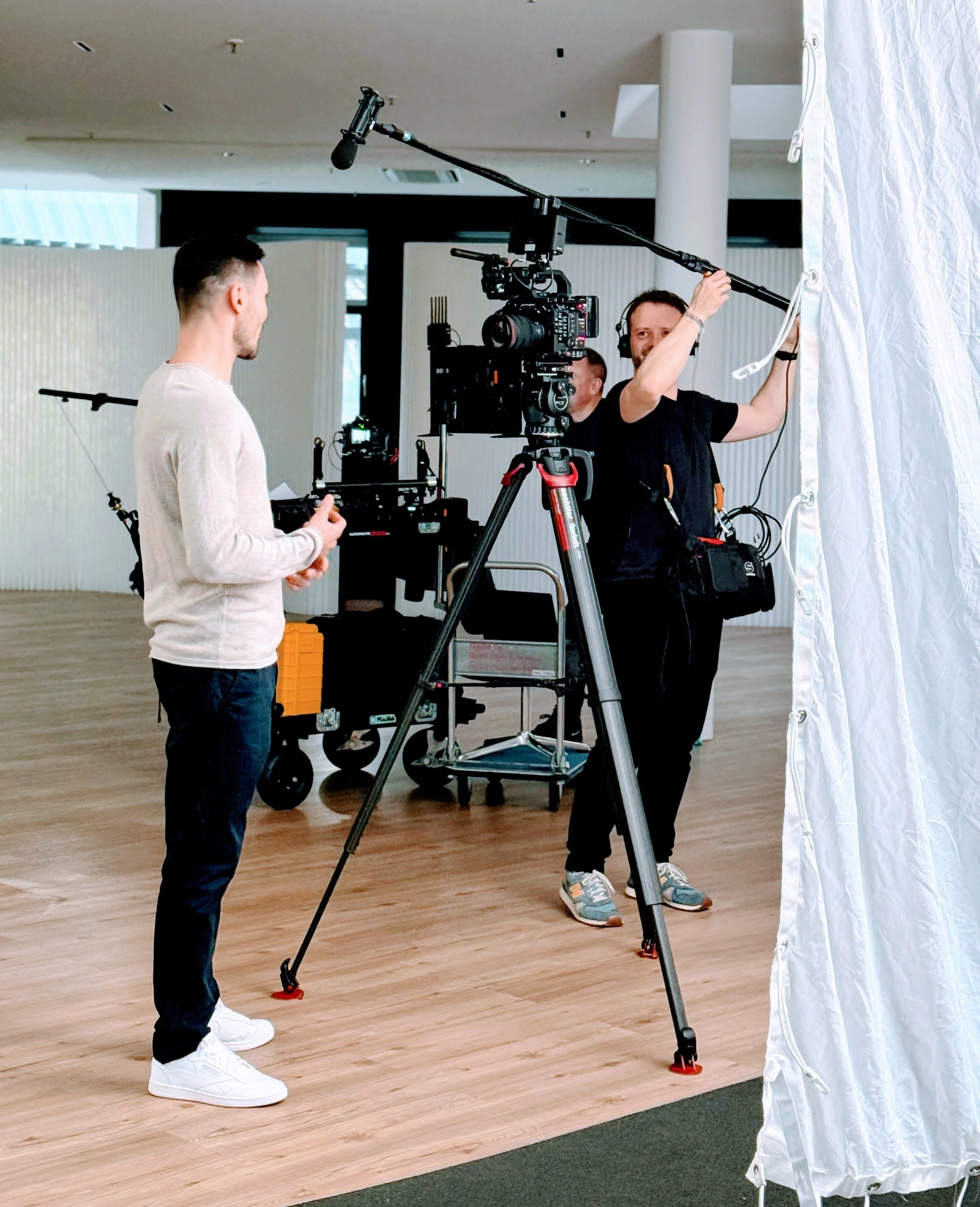 Filmcrew bei der Arbeit, eine Person bedient eine Kamera auf einem Stativ, eine weitere Person bereitet einen weißen Vorhang für die Aufnahme vor, im Hintergrund sind leere Wände und ein Holzboden.