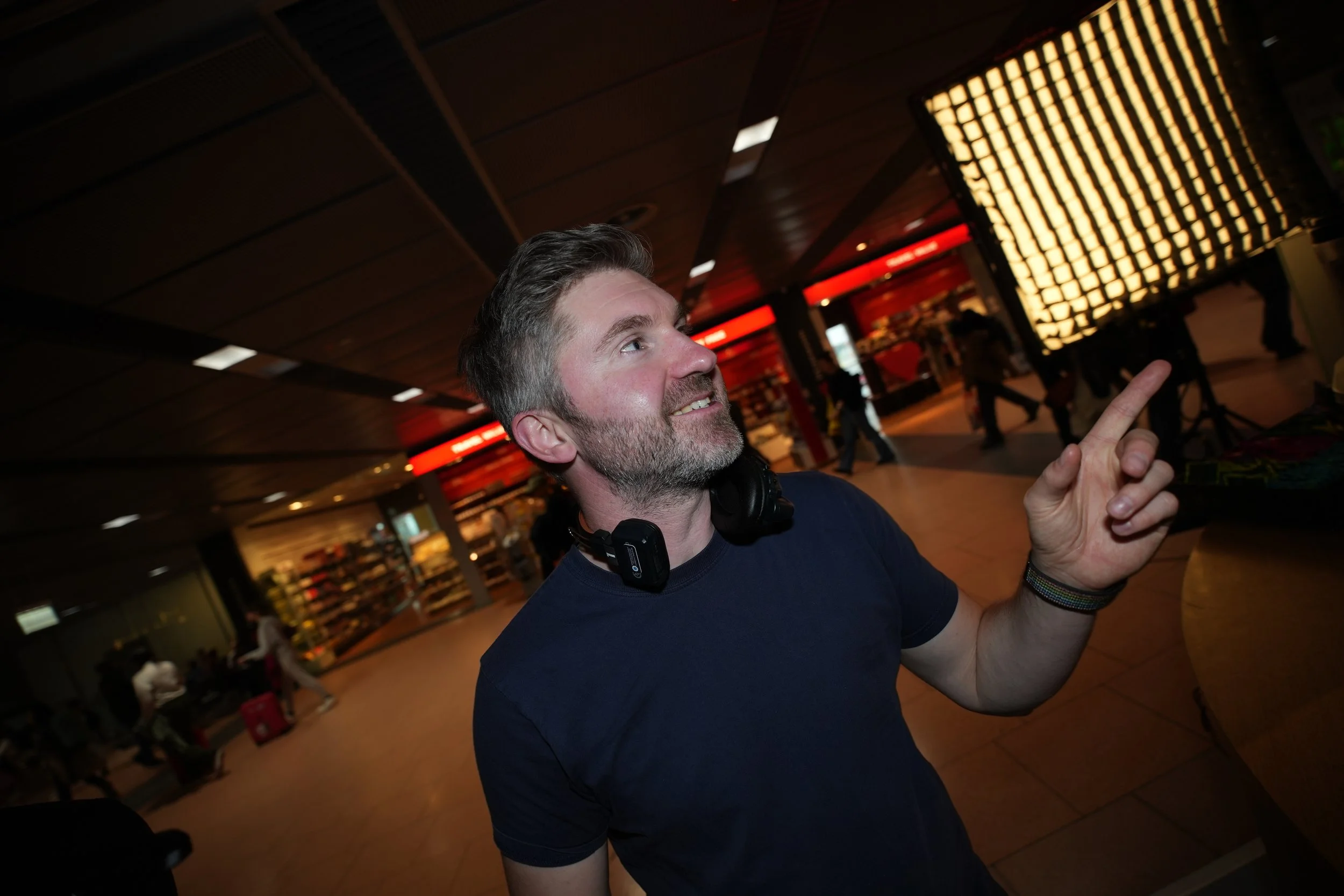 Ein lachender Mann mit Kopfhörern um den Hals zeigt in einem Flughafengebäude mit Rückenleuchten und anderen Reisenden auf eine Lichtinstallation, vermutlich in einem Wartebereich.
