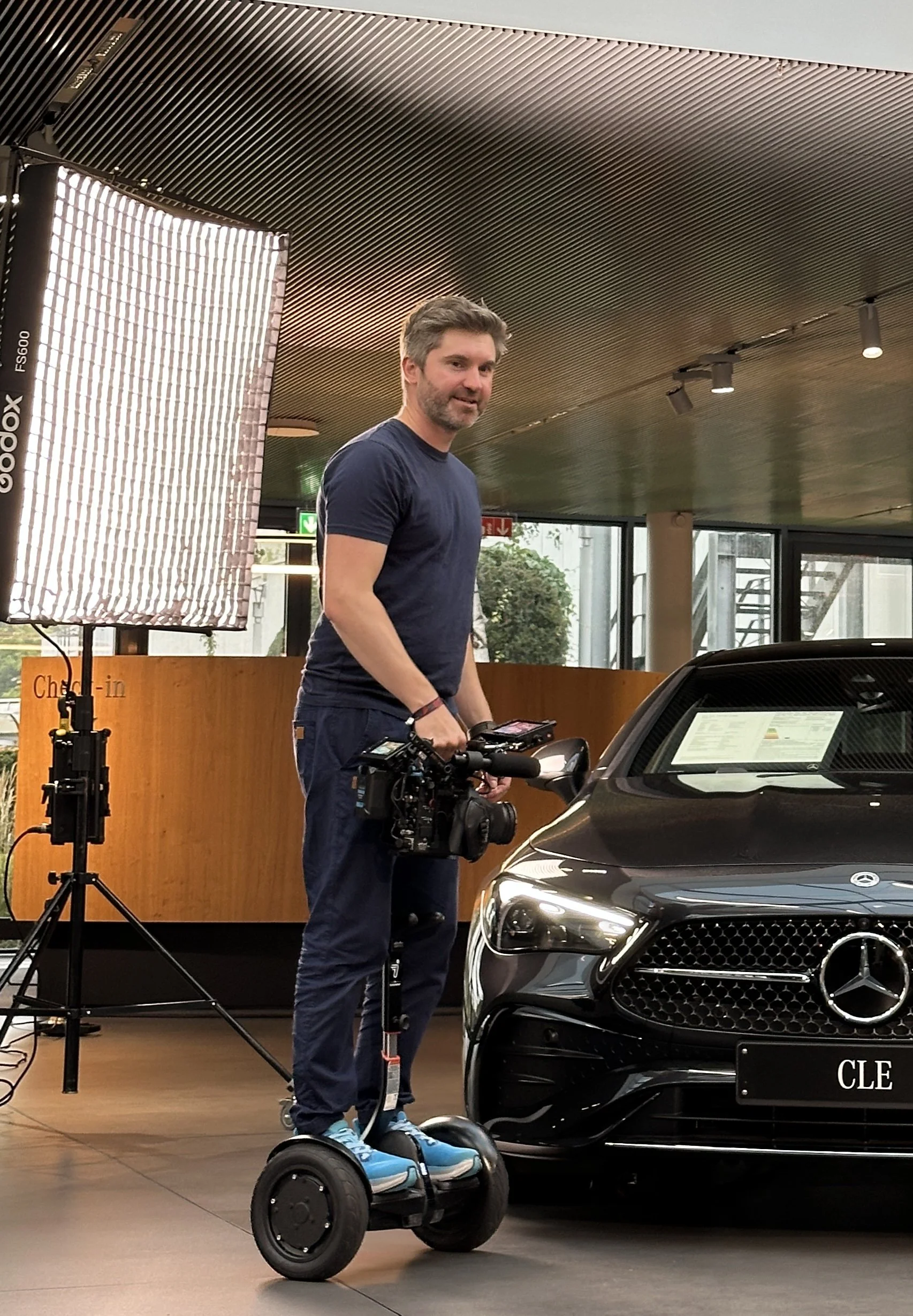 Mann auf einem Segway neben einem schwarzen Mercedes-Benz Auto in einem Autohaus, während einer Videoaufnahme.