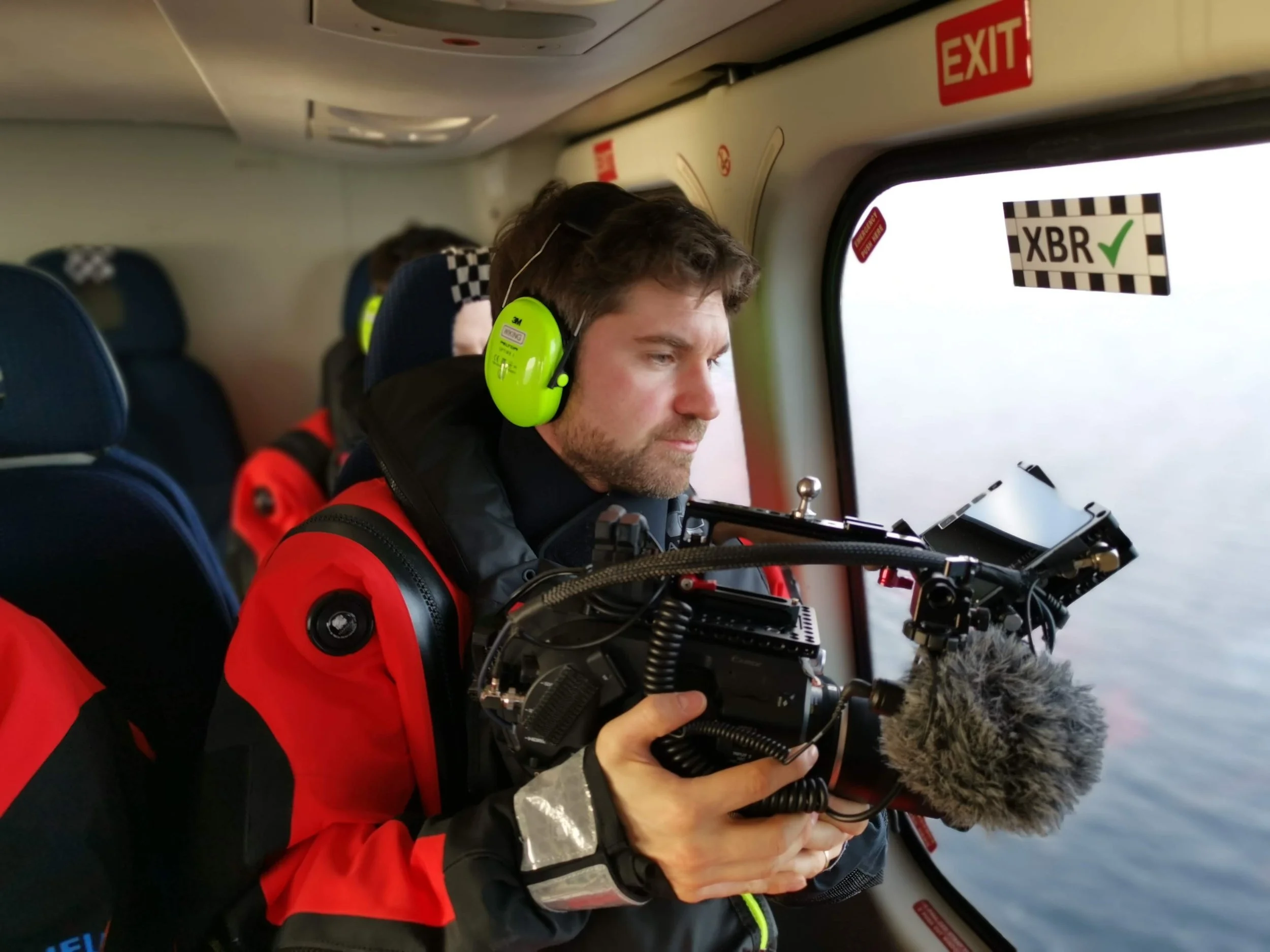 Ein Mann mit Kopfhörern und Kamera sitzt im Flugzeug, während er aus dem Fenster schaut.