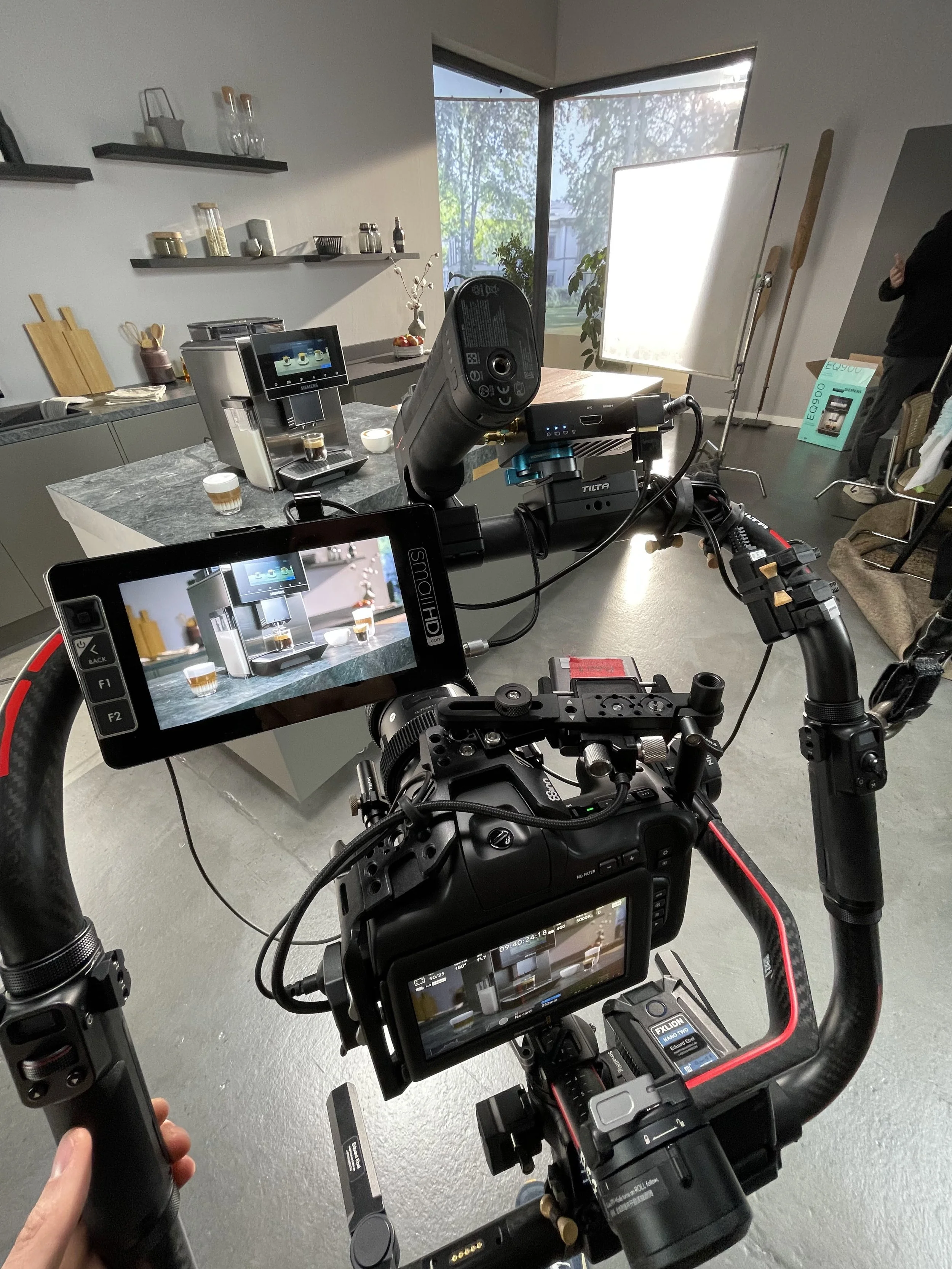 Aufnahmegerät filmt eine Kaffeemaschine auf einem Tisch in einem modern eingerichteten Raum, mit großen Fenstern im Hintergrund und einem Bildschirm, der das Video zeigt.