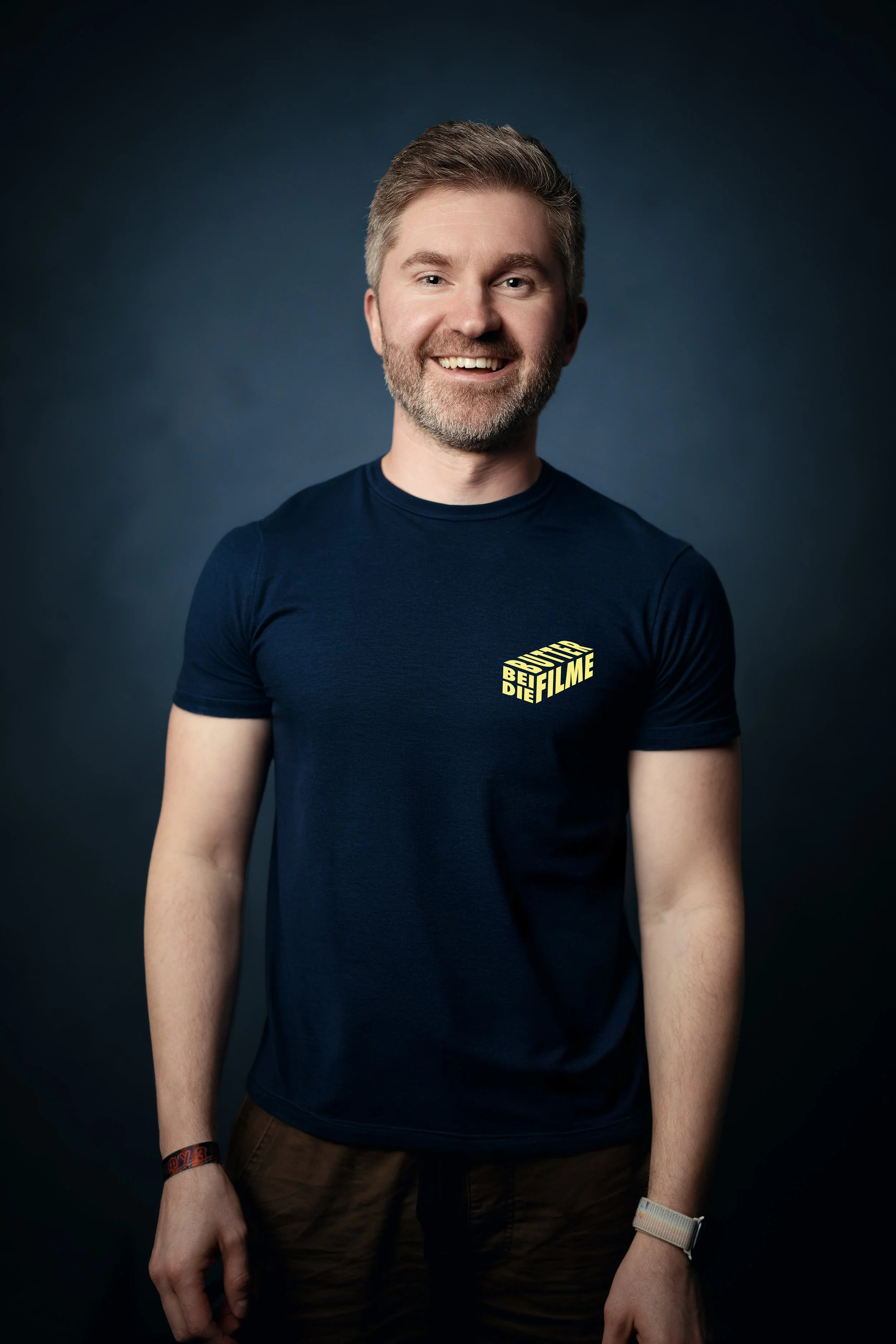 Ein lachender Mann mit Bart und grauem Haar vor dunklem Hintergrund, trägt ein dunkelblaues T-Shirt mit gelbem Logo, das 'BLETZ DEr FILME' lautet.