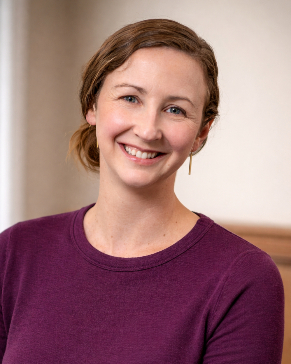 A woman with short light brown hair, wearing a purple shirt and gold earrings, smiling at the camera.