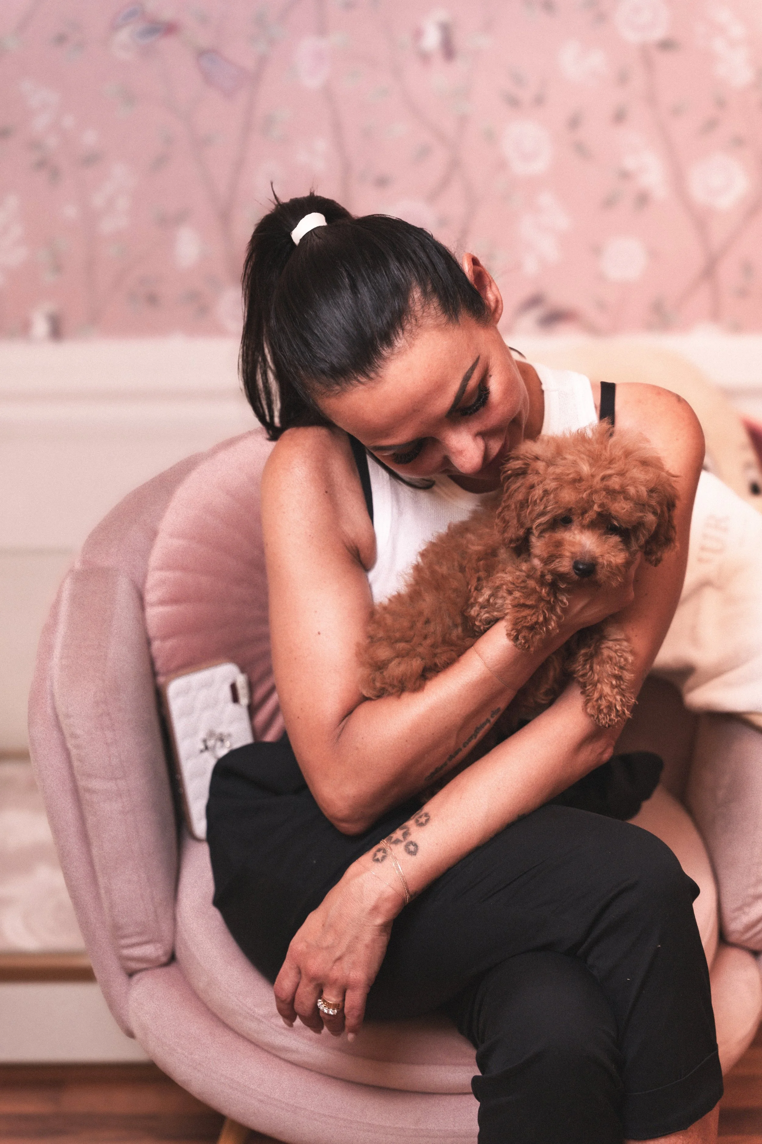 Una donna con capelli raccolti in una coda, vestita di bianco e nero, abbraccia un piccolo cucciolo di cane di pelo riccio marrone, seduta su una poltrona rosa in un ambiente domestico.
