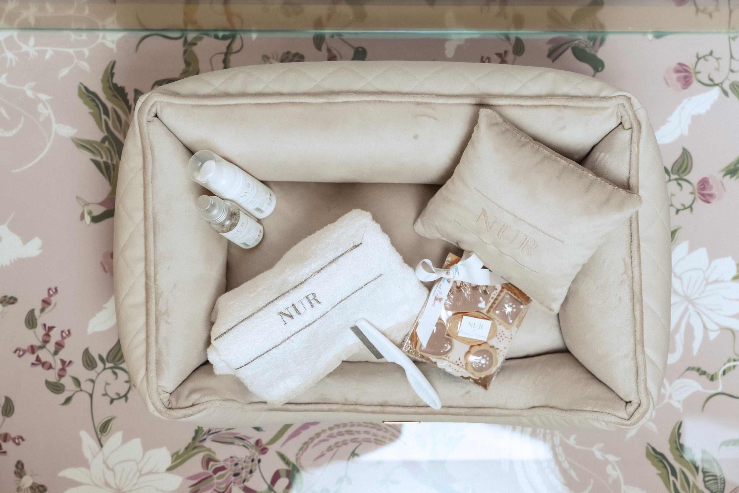 Set di prodotti di bellezza su una scatola rivestita di tessuto beige, posizionata su una superficie con motivo floreale rosa, verde e bianco.