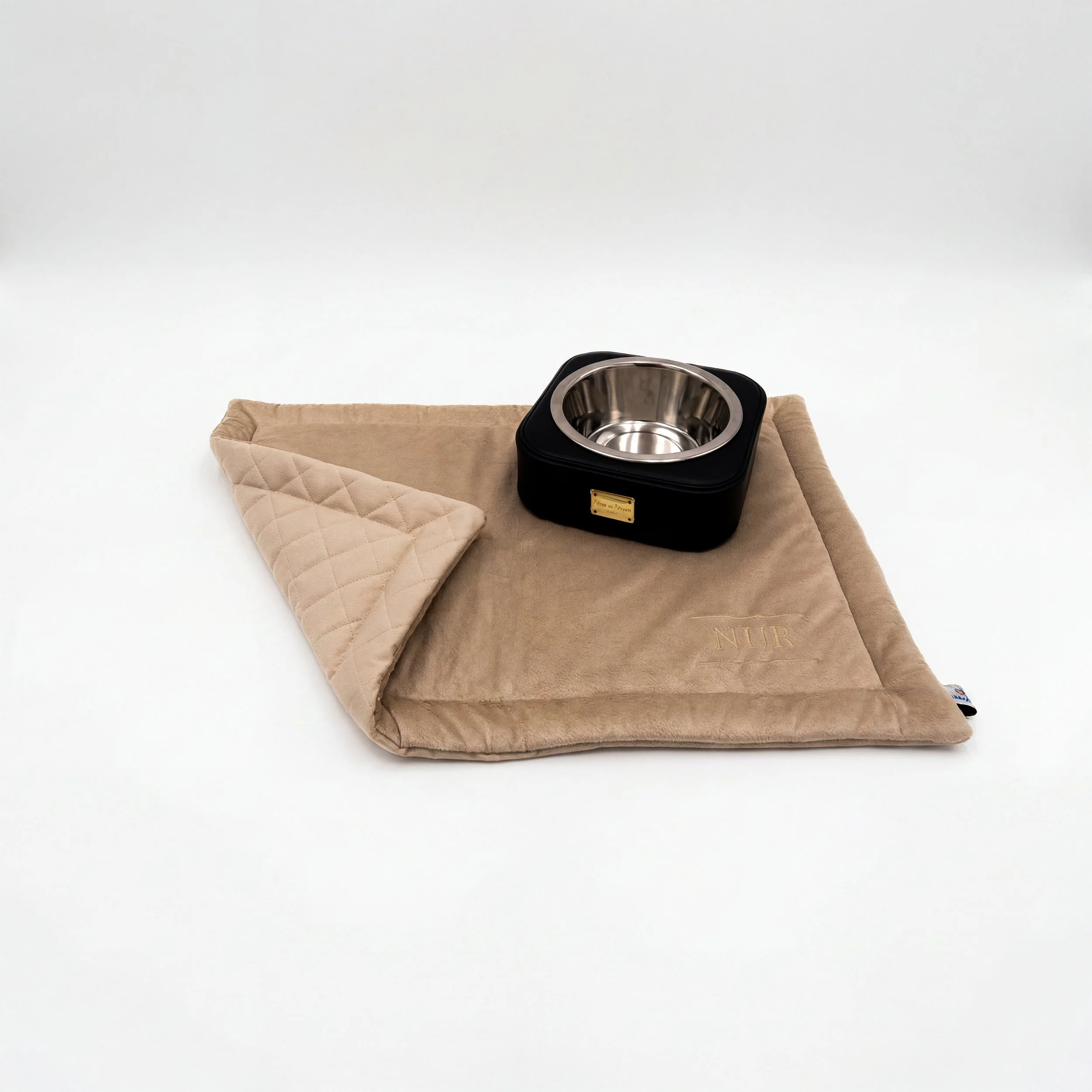 Un divano beige con un cuscino abbinato su una superficie bianca, davanti a un recipiente in acciaio inox su una scatola nera con etichetta dorata.