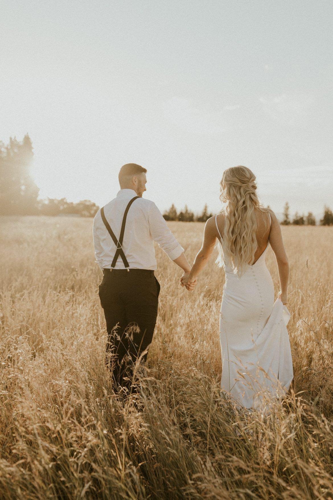 A couple dressed in wedding attire walking hand in hand through a field of tall grass at sunset.