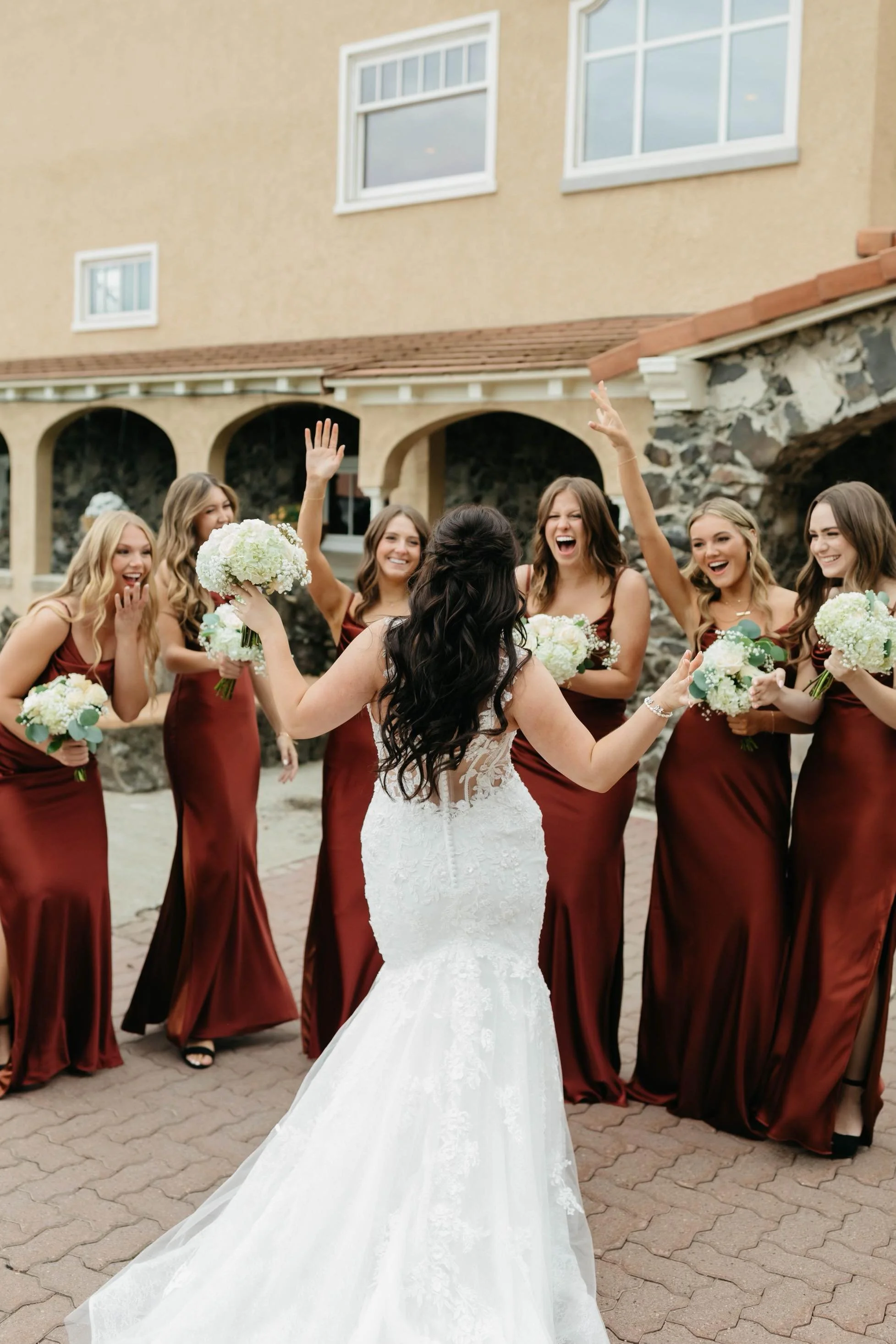 A bride in a white lace wedding dress holding a bouquet and celebrating with her bridesmaids dressed in matching maroon gowns holding bouquets outside a building with stone and stucco facade.