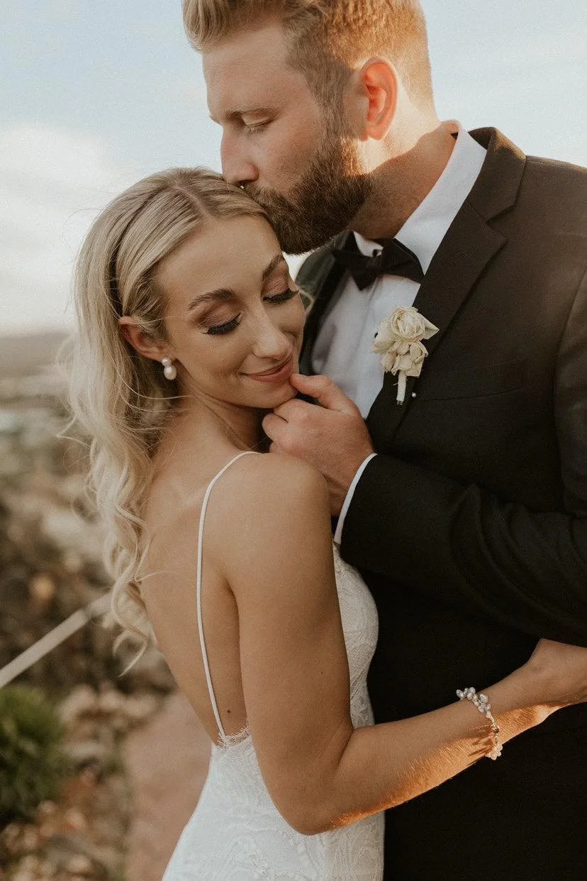 A newlywed couple sharing an intimate moment, with the groom kissing the bride on her forehead, outdoors during sunset.