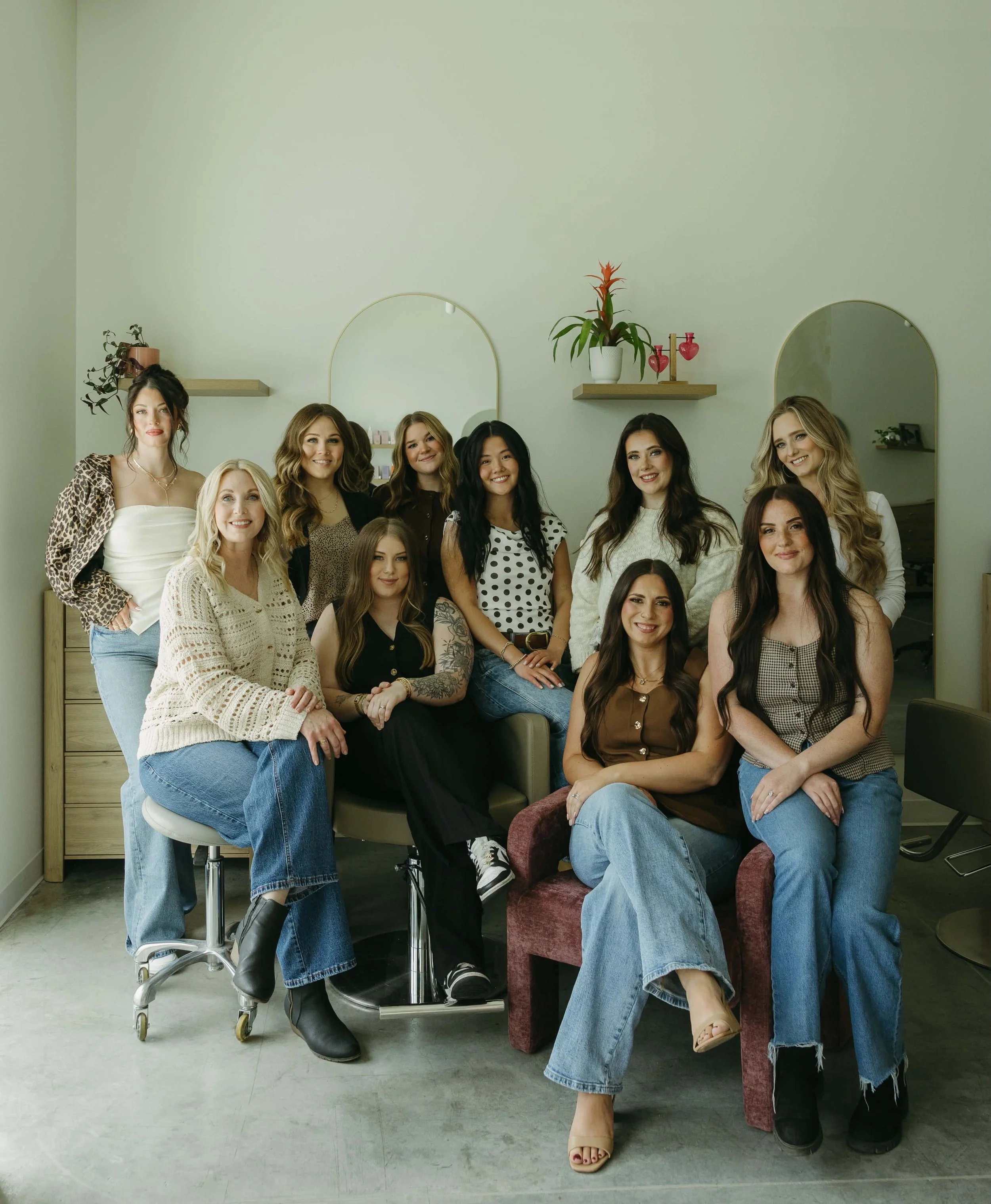 A group of eleven women posing together indoors in front of mirrors and light green walls with shelves and plants, some standing and some seated.