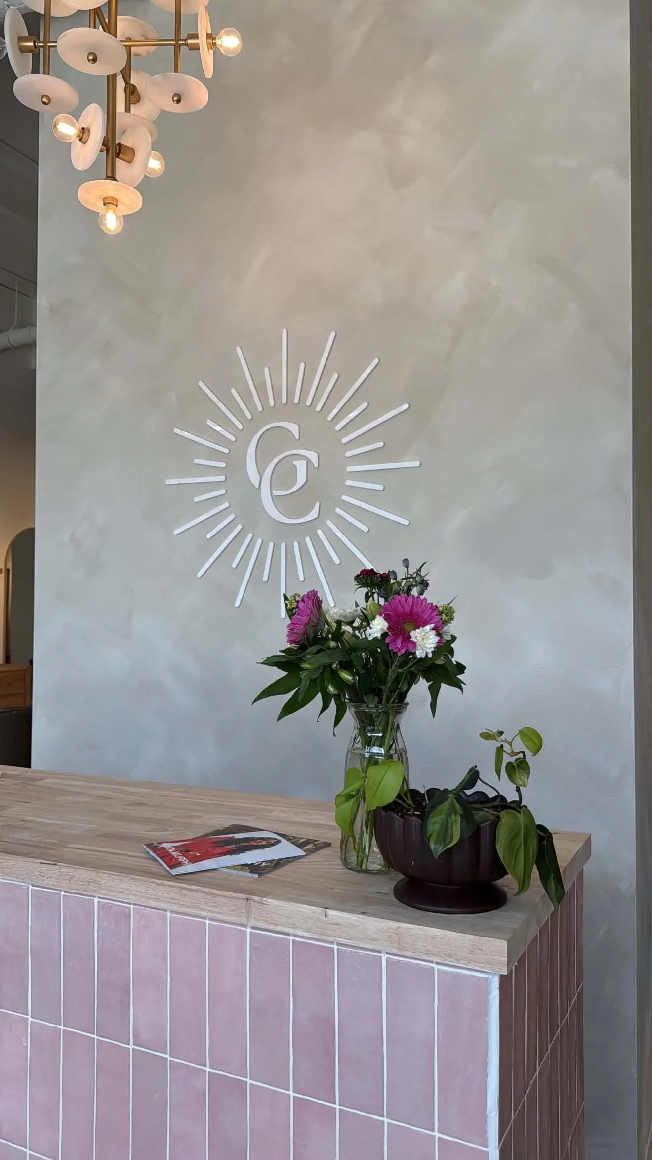 A reception desk with a pink tiled front, a clear vase with pink and white flowers, and a black planter with green plants. A wall with a logo consisting of the letters 'G' and 'C' inside a sunburst design is visible behind the desk. A modern chandelier with multiple white and gold elements hangs from the ceiling.