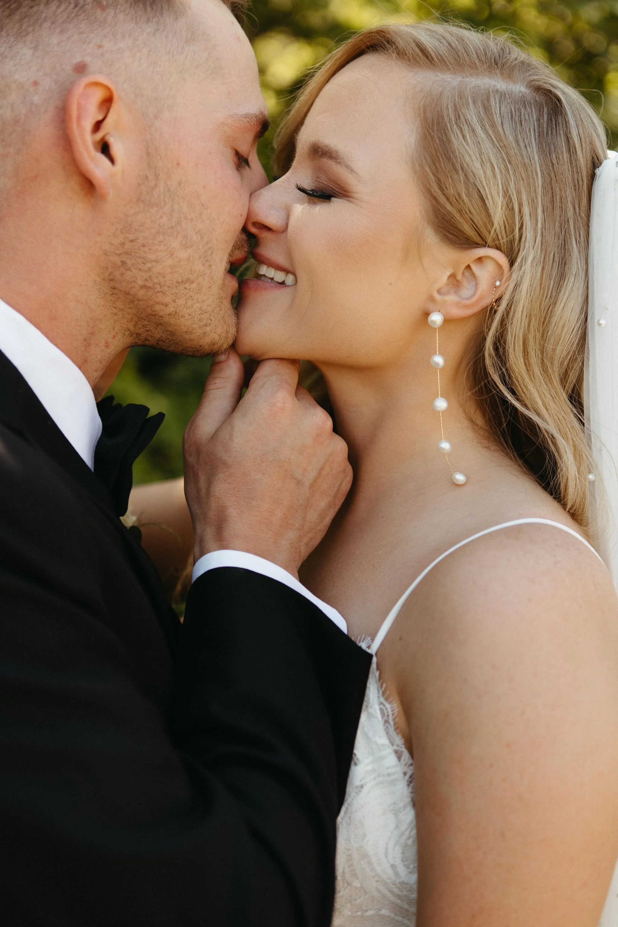 A close-up of a wedding couple sharing a kiss outdoors, with the bride wearing pearl earrings and a white lace dress, and the groom in a black tuxedo with a bow tie.
